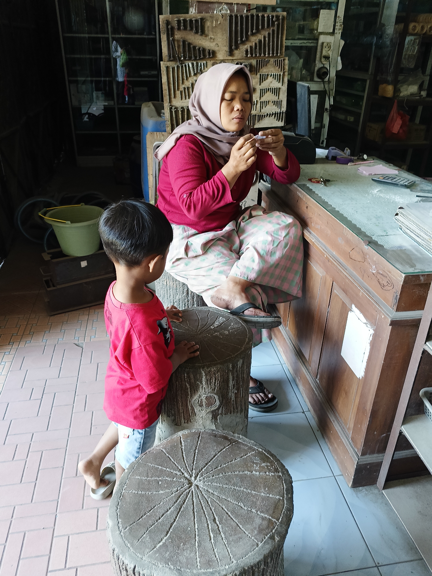 A woman wearing a hijab and a pink shirt sits at a wooden counter in Sukorini, Indonesia, meticulously working with small objects in her hands. Beside her, a young boy in a red shirt and shorts stands on what appears to be a textured, log-shaped stool, observing the woman. The environment suggests a workshop or shop, with shelves filled with various items and tools visible in the background. The counter has a glass top, and a calculator and other small objects are scattered on its surface. The floor is tiled.