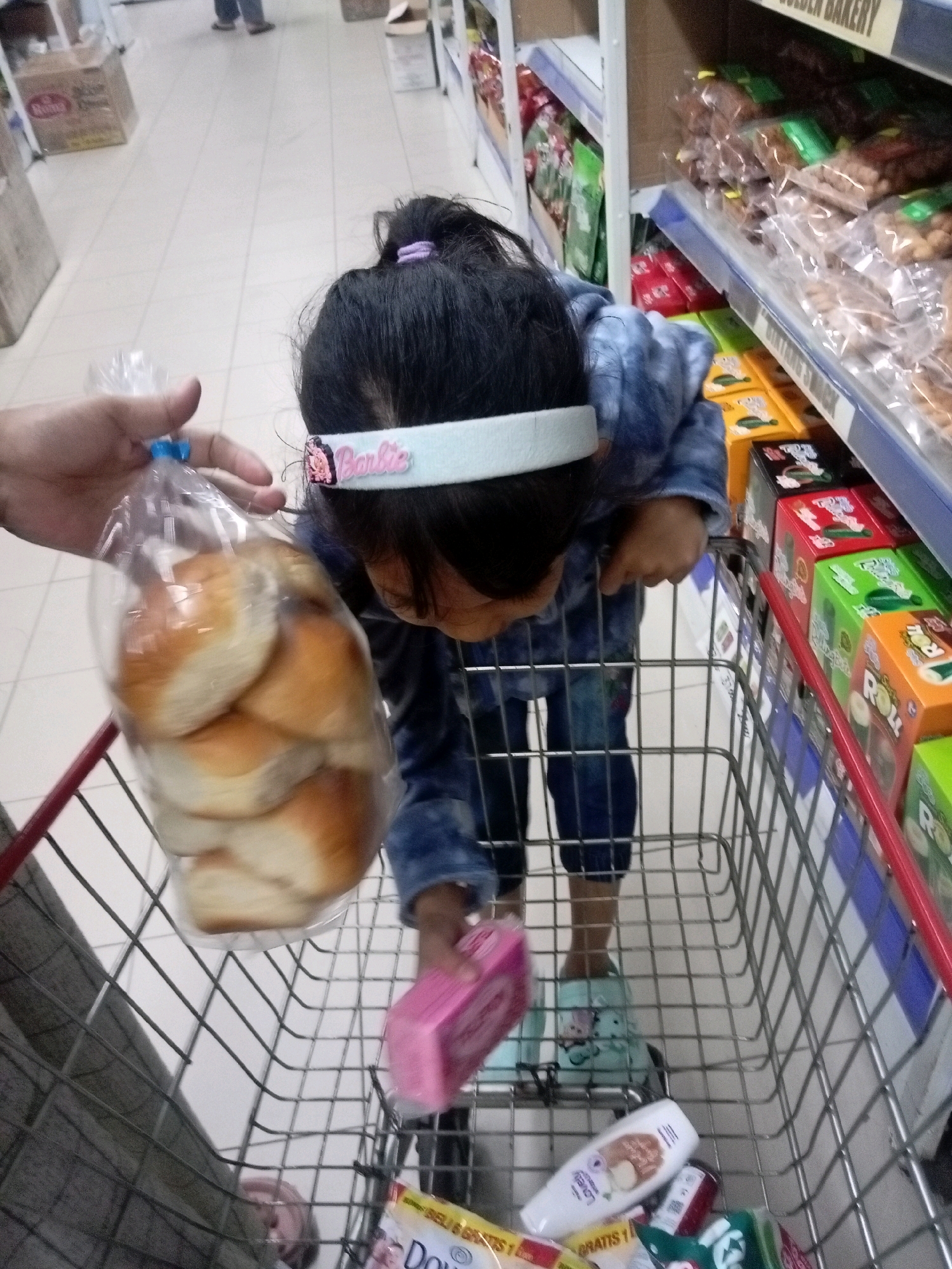 A young child, wearing a blue patterned long-sleeved shirt and blue jeans, stands in a shopping cart in a retail store. The child has dark hair, pulled back with a purple elastic band and a white headband with "Barbie" printed on it. The child is holding a clear plastic bag filled with bread rolls and is reaching for a pink box. The shopping cart contains other items, including a bottle of what appears to be lotion, a package of tissues, and several boxes of snacks. The store shelves are visible in the background, stocked with various food items. The floor is tiled, and the overall lighting suggests an indoor commercial environment. Based on the visible products and store layout, this scene is likely in a supermarket or minimarket in Batam, Indonesia.