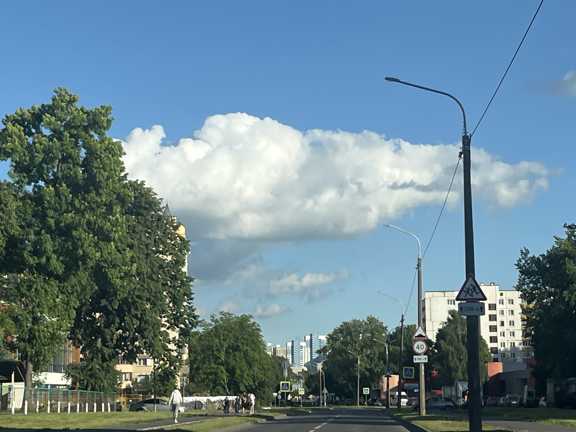 The image depicts a street scene in Minsk, Belarus, under a bright, sunny sky with scattered white clouds. The time of day appears to be late morning or early afternoon. The weather is clear and pleasant.
The foreground shows a paved road with markings and a grassy verge. On the left side of the road, a large, leafy green tree dominates the view, with a building partially visible behind it. A fence runs along the edge of the property. A single person is walking on the sidewalk.
Further down the street, there are more trees and buildings, including a tall, modern apartment building. Several streetlights line the road. Traffic signs are visible, including a triangular sign with a warning symbol of people crossing and text that appears to read "200 m". Another sign indicates a speed limit of 40. A yellow pedestrian crossing sign is also present.
In the middle ground, a group of people are walking together on the sidewalk, and a few vehicles are parked or driving. In the distance, several taller buildings, possibly office or residential complexes, can be seen against the sky. The overall impression is a typical urban environment during a pleasant day.