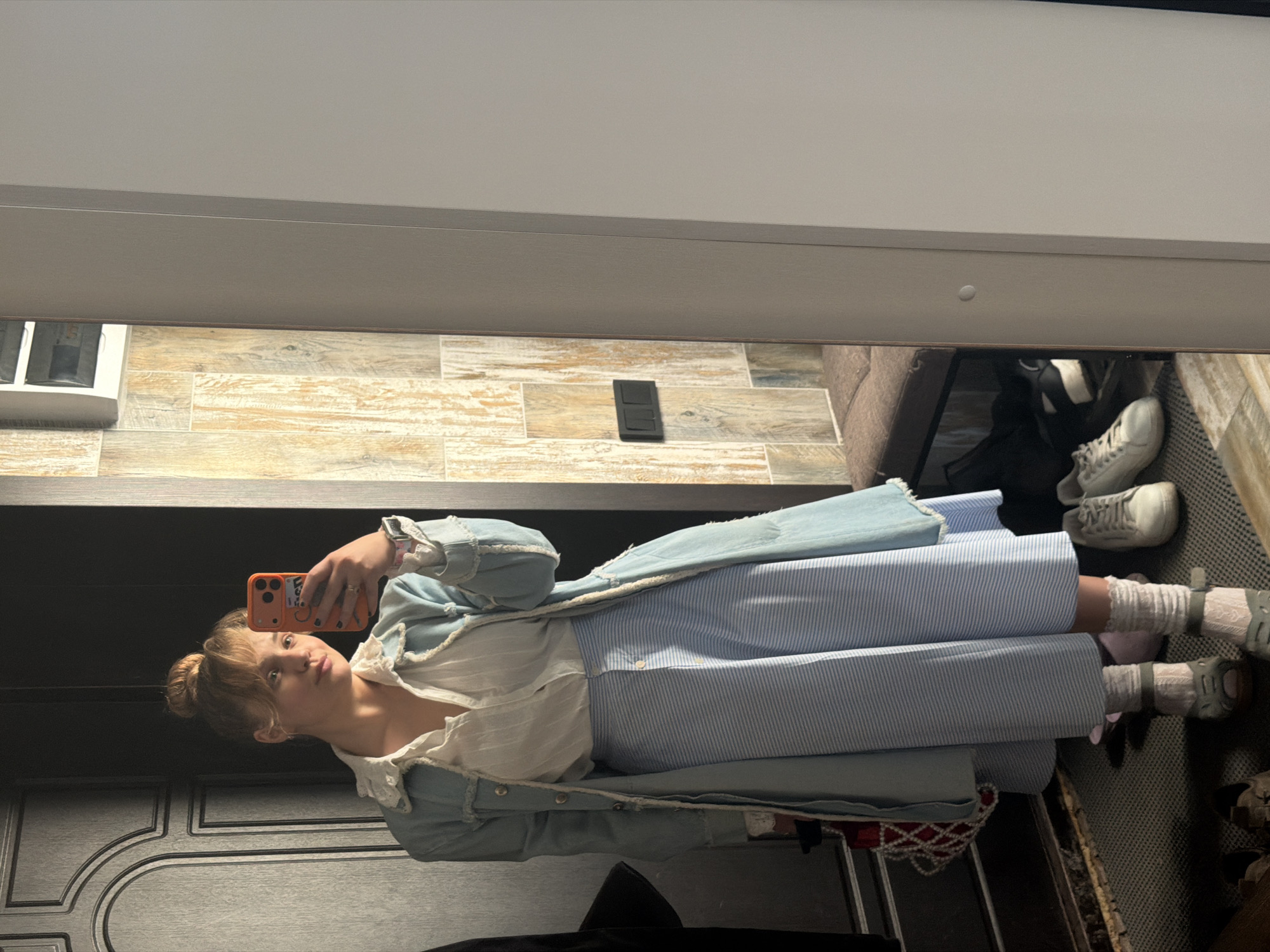 This indoor mirror selfie, taken in Большая Шильна, Russia, features a young woman posing in front of a mirror. She has light-colored hair styled in a bun, with some strands framing her face, and is looking directly at the camera with a slightly pouty expression.
She is dressed in a light blue, denim-style long coat with distressed edges, worn over a white blouse featuring a ruffled collar and front. Below the blouse, she wears a light blue and white striped long skirt. On her feet, she has light-colored socks, possibly pink or white, paired with open-toed, light green or grey sandals. She holds an orange smartphone in her right hand, using it to capture the reflection. A smartwatch is visible on her left wrist.
The reflected background reveals an interior setting. The wall behind her features light-toned wooden plank or tile paneling, with a dark electrical outlet mounted on it. To the right, part of a beige or brown couch and a pair of white sneakers on a patterned mat are visible. To the left, a dark door with ornate molding can be seen. The ceiling is a plain light gray. The lighting suggests an indoor setting, possibly artificial. No discernible text is visible on any objects in the image.