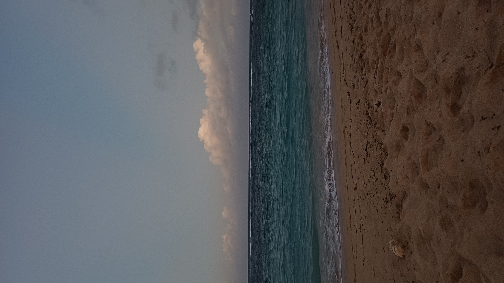 The image shows a beach scene in Kuta Selatan, Indonesia. The view is vertically oriented, with the sky occupying the left portion of the frame, and the beach taking up the right. A narrow strip of ocean separates the two. The sky is a muted blue-gray color, with a line of puffy white clouds near the horizon. The ocean is a mix of dark blue and turquoise, with small white waves lapping the shore. The sand is a dark tan color and appears coarse, with many footprints visible. A small, light-colored object, possibly a rock or shell, sits on the beach near the bottom of the frame. There are no people visible.