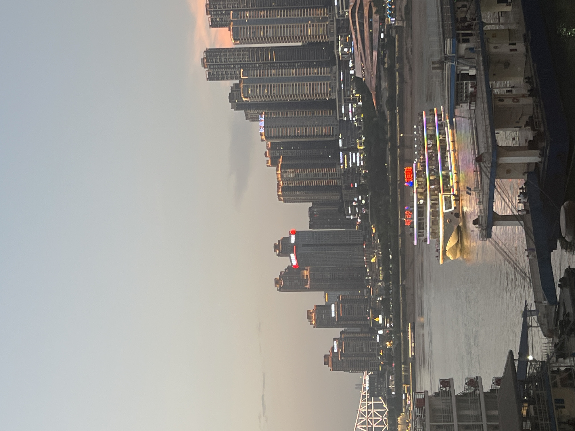 The image depicts a wide river flowing through a city at dusk. On the right bank of the river, a dense collection of modern skyscrapers with illuminated windows and signs dominates the skyline. The buildings are of varying heights, creating a staggered effect. A bridge with illuminated white geometric structures is visible on the left side of the image, spanning the river.
In the foreground, on the river, a brightly lit cruise ship with multiple decks is moving from left to right. The ship is adorned with colorful lights that reflect on the water. Several other vessels, including what appear to be barges or smaller boats, are docked or moored along the left bank of the river, with some people visible on their decks. The sky is a soft gradient of grey and pale orange, suggesting the late evening or twilight hours. The water is calm, with subtle ripples reflecting the city lights. The overall scene captures a vibrant urban waterfront environment.