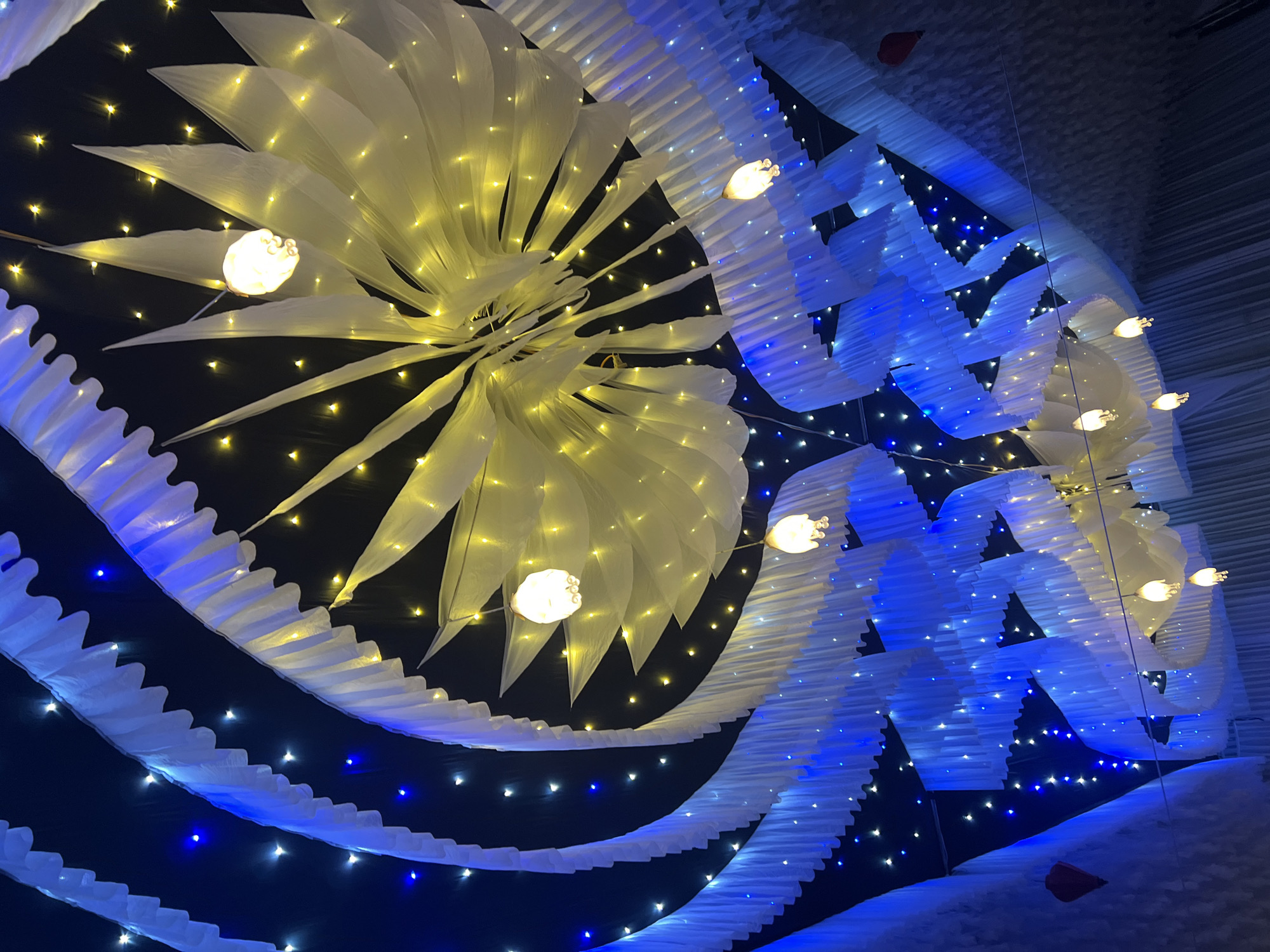 The image displays an elaborate, illuminated decorative installation, likely part of an indoor event or art exhibit, set against a dark background. The dominant feature is a large, central arrangement in the upper left, resembling a radiant starburst or a blooming flower, fashioned from numerous translucent white or off-white fabric petals. This feature is heavily adorned with warm yellow string lights, creating a soft, glowing effect, and several larger, spherical white light bulbs are integrated within it.
Flowing across the right and bottom portions of the frame are undulating, wave-like structures made from accordion-folded white material. These elements are illuminated by cool blue and white lights, creating a dynamic contrast with the yellow hues of the central piece. Small, twinkling lights in both yellow and blue are scattered across the dark background, enhancing the magical and ethereal atmosphere, reminiscent of a starry night or shimmering water.
No people or activities are visible, placing sole emphasis on the intricate decorations. The scene evokes a sense of wonder and festivity, potentially representing a winter theme or a fantasy landscape. The lighting suggests it is either night or a purposefully darkened environment to highlight the illuminations. This striking display is located in Bình An, Vietnam. There is no visible text in the image.