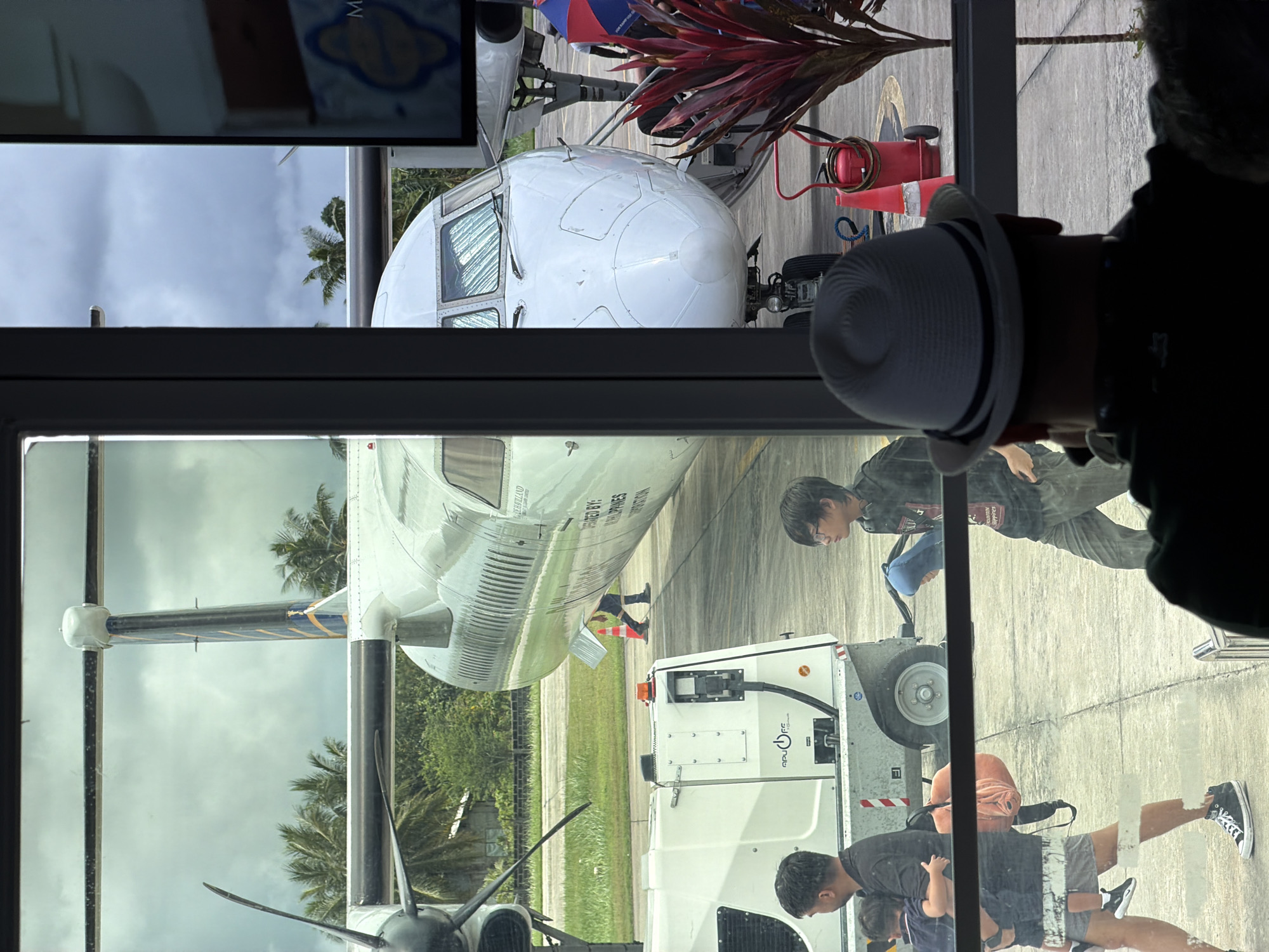 The image shows an ATR-72 aircraft parked at a gate in Del Carmen, Philippines, seen through a glass window. The plane's nose and part of the fuselage are visible outside. A ground support vehicle is also visible. Several people are walking around outside, including one person wearing a hat and another with glasses. A red fire extinguisher on wheels and red leafy plant. Palm trees are visible in the background. The glass window reflects the inside of what looks like a waiting area.