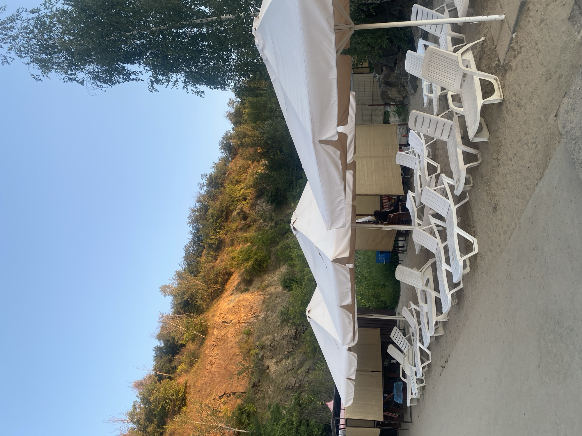 The image depicts a beach area with several white plastic lounge chairs lined up on the sand. Large white umbrellas are erected over some of the chairs and covered seating areas. In the background, a steep, rocky hillside covered in trees rises towards a clear blue sky. The sun is casting a warm glow on the hillside, indicating it is likely late afternoon or early evening. Some covered structures resembling cabanas or small gazebos are visible behind the lounge chairs, with what appears to be seating and tables inside. A few people can be vaguely seen within these structures. A tall birch tree stands on the right side of the image. The scene suggests a leisure or resort setting, possibly a beach club or a public beach with amenities. This location appears to be a beach along the Dnipro River in the city of Dnipro, Ukraine.