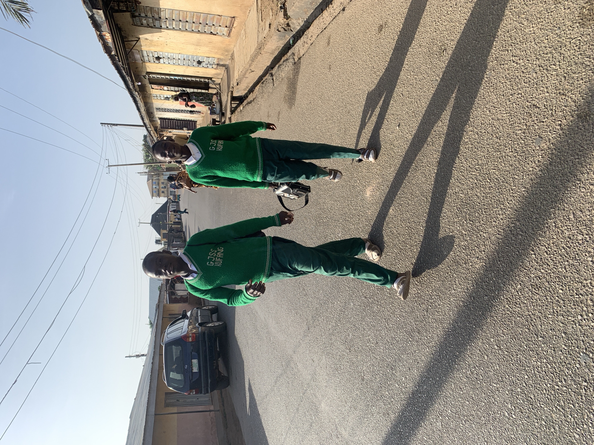 Two young men are standing on a paved street in Jos, Nigeria. They are wearing matching green school uniforms consisting of sweaters with the inscription "G.J.S.S. KUFANG" and dark pants. The shadows they cast suggest a sunny day. Behind them are buildings and parked cars. Overhead are telephone poles and wires.