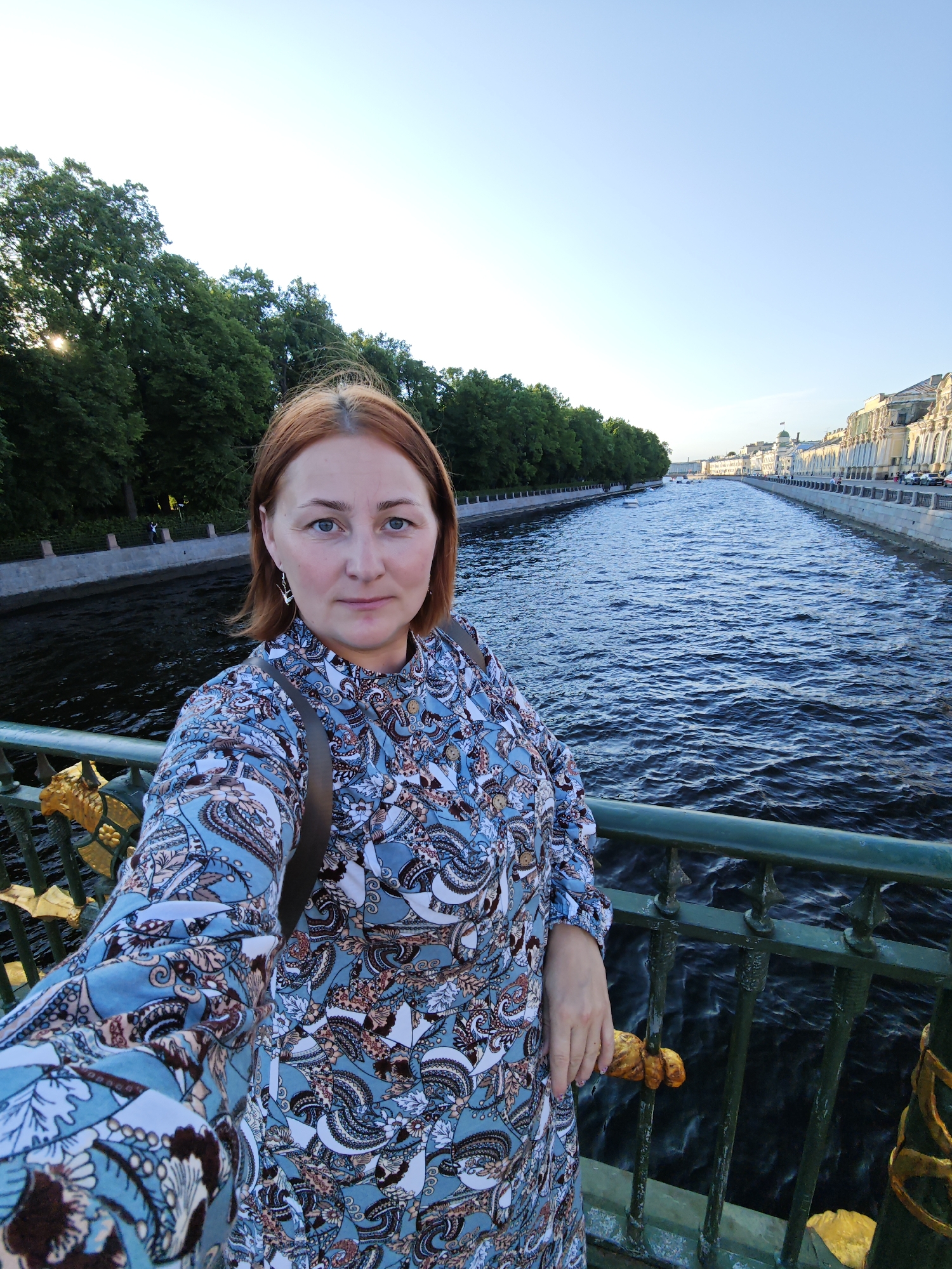 A woman with auburn hair is taking a selfie on a bridge overlooking a wide canal in Saint Petersburg, Russia. She is wearing a blue and white patterned dress and a brown backpack. The canal is dark blue with ripples on its surface and is lined with trees on the left and ornate buildings on the right. The sky is clear and pale blue. The bridge has a green railing with decorative golden elements.