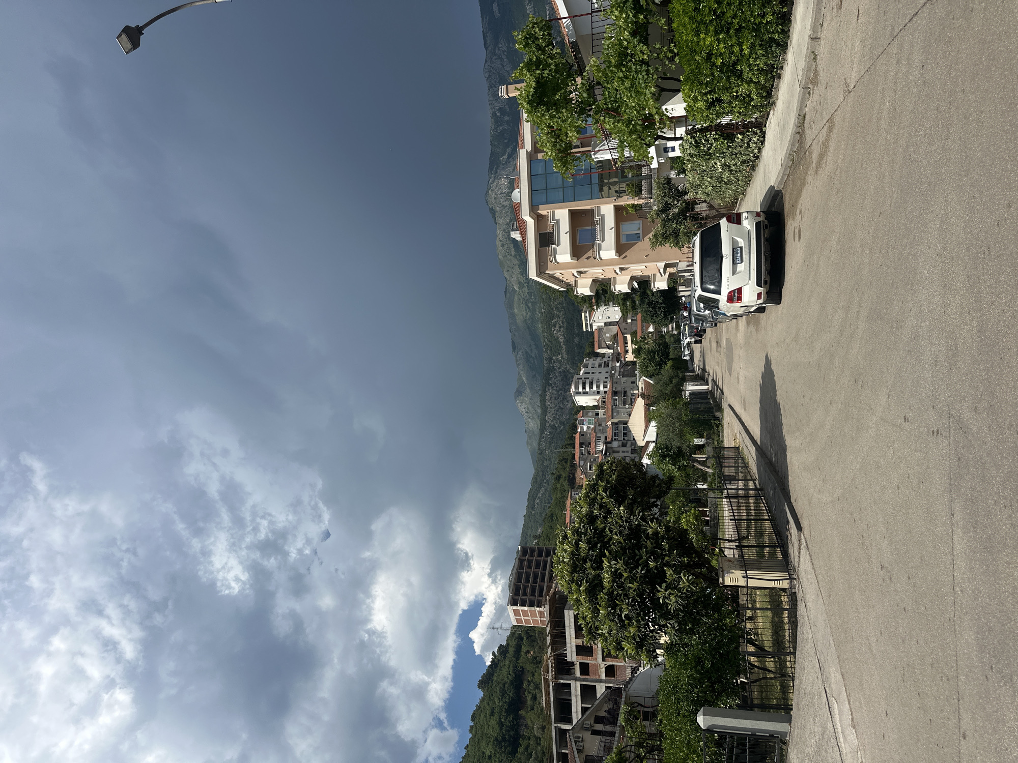 The image depicts a street scene in Budva, Montenegro. The perspective is from the side of the road, looking upwards and across a town that slopes towards a mountainous backdrop. The sky is dramatic, with a stark division between dark, stormy clouds on the left and bright, sunlit areas on the right, suggesting a passing storm or a strong contrast in weather patterns.
In the foreground, the asphalt road curves gently to the right. Along the left side of the road, there's a sidewalk and a low metal fence, beyond which are lush green trees and the lower levels of buildings. A white SUV is parked on the right side of the street, casting a shadow. Further down the street, more cars are visible, and the architecture consists of modern buildings with multiple stories, some appearing residential, others commercial. One building, possibly under construction, shows exposed concrete framework.
The middle ground is dominated by the town itself, nestled against the green, mountainous terrain. The buildings are a mix of colors, primarily beige, white, and some with modern glass facades. The mountains in the background are covered in vegetation, and the scale suggests a significant elevation change.
In the upper left corner, a street lamp is visible against the turbulent sky. The overall impression is one of a coastal town experiencing varied weather conditions, with a blend of urban development and natural landscape. The time of day appears to be late afternoon or early evening, given the angle of the sun and the shadows. No people are clearly visible, and the focus is on the urban and natural environment.