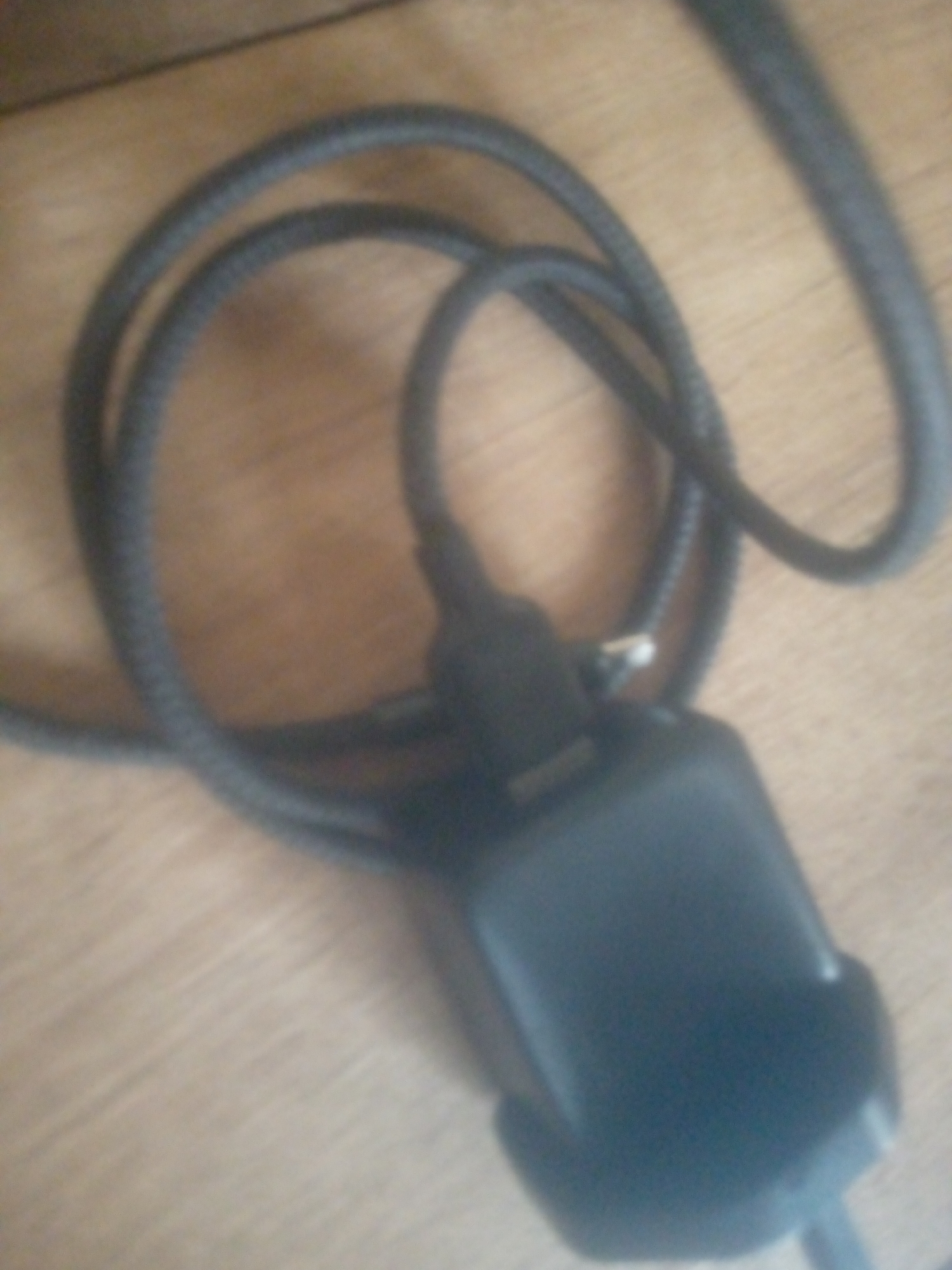 A black coiled cable with a black plug is pictured on a wooden surface. The cable is looped and draped across the surface. The plug is rectangular and has a metal connector visible. The image is blurry and out of focus, making it difficult to discern fine details. The lighting is dim.