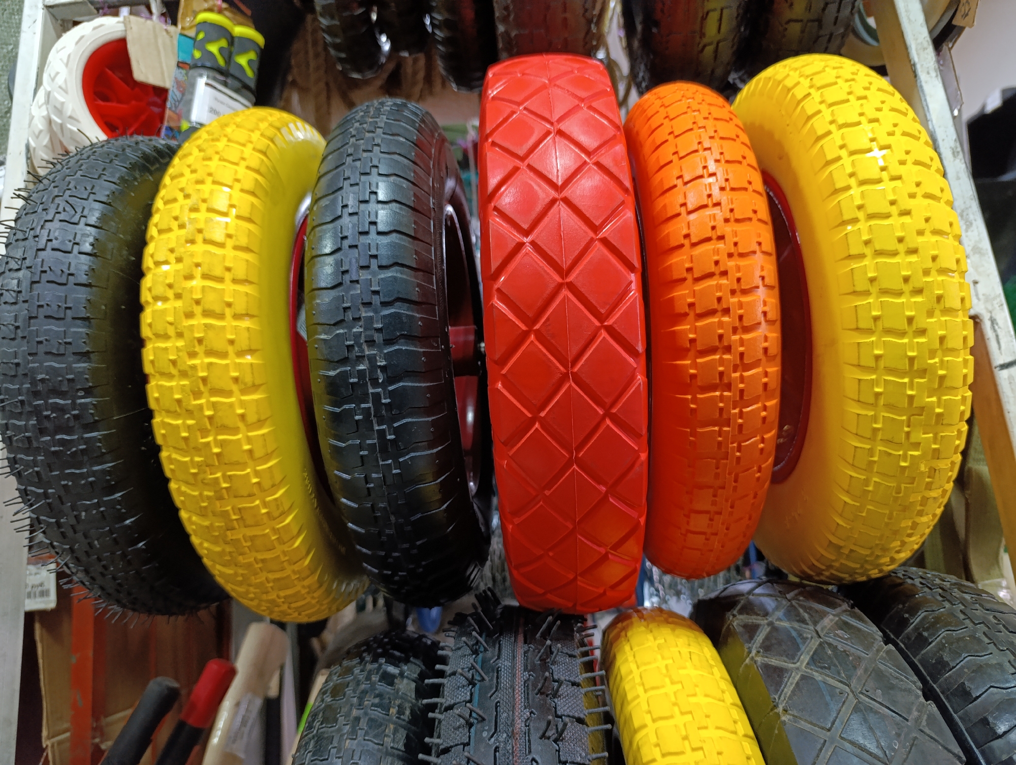 This media file captures a close-up view of a vibrant collection of small wheels, likely intended for hand trucks, wheelbarrows, or similar utility equipment. The items are neatly arranged on what appears to be a display shelf or storage rack in an indoor setting, suggestive of a hardware store, workshop, or merchandise area.
The wheels are a prominent feature, showcasing a striking array of colors: bright yellow, bold red, and vivid orange, interspersed with standard black. Each wheel displays a distinct tread pattern; for instance, the central red wheel features a geometric diamond-shaped pattern, while the yellow and orange wheels have more uniform, rectangular block treads. Some of the black tires exhibit small rubber "hairs" or nubs, indicating they are new. Many of the wheels have visible hubs, typically in contrasting colors like red or black.
In the background, other indistinct objects can be seen, including hints of tool handles with red and black grips. On a white label attached to a packaged item partially visible in the upper left section of the image, the number "200" is clearly discernible, possibly representing a price or stock identifier. The lighting is consistent with indoor artificial sources, creating subtle reflections on the glossy surfaces of the colored wheels. No activity is taking place; the image is a still life focusing on the products. The scene is located in Chaikovsky, Russia.