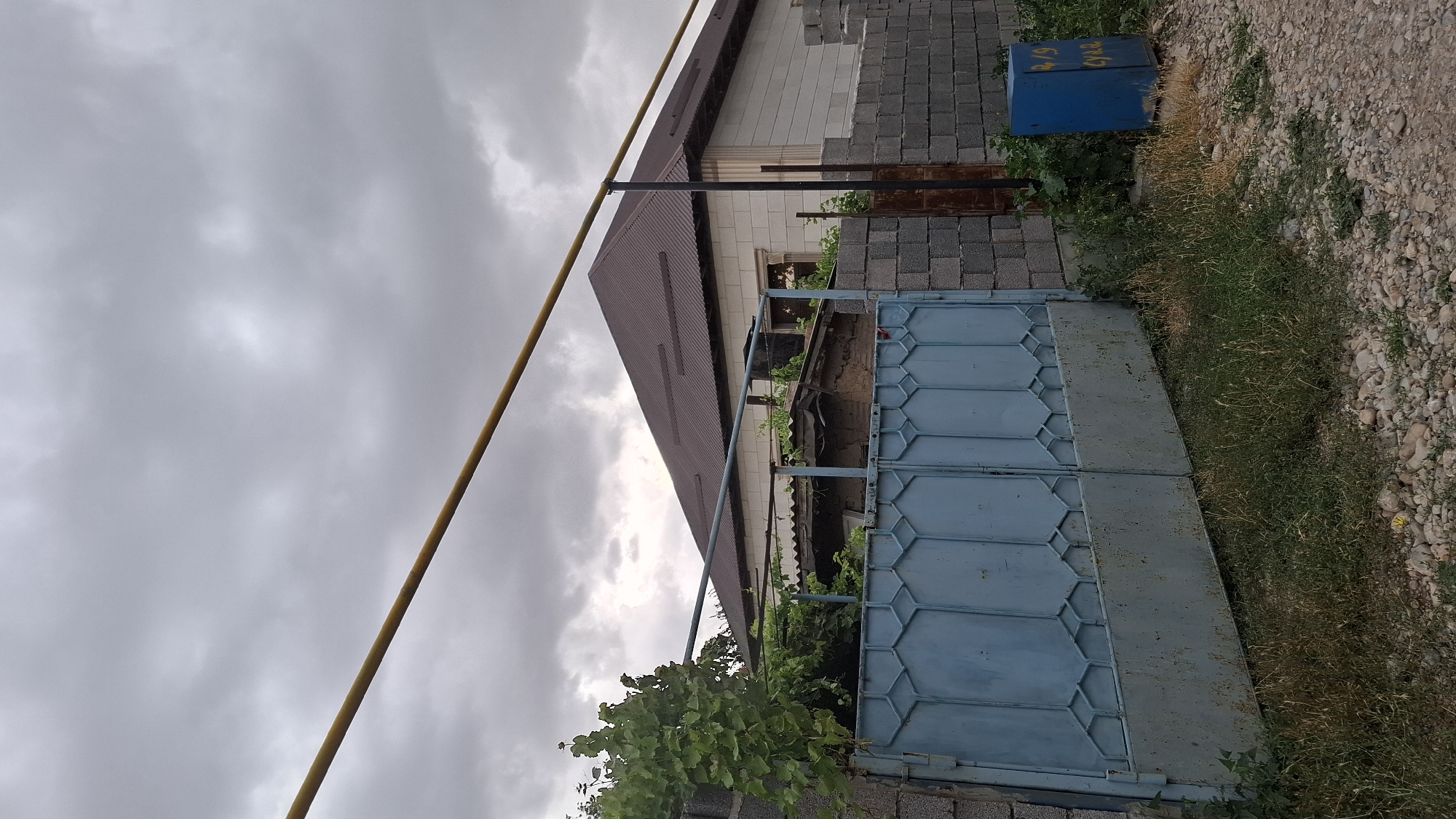 The image depicts the exterior of a residential property in Шұбарсу, Kazakhstan. A blue, corrugated metal gate with a decorative geometric pattern is partially open on the left side of the frame. Behind the gate, a low-slung, weathered structure with a corrugated roof is visible, overgrown with green foliage. To the right of the gate, a brick wall forms the boundary of the property. Beyond the gate and the foreground structure, a larger house with a dark, sloped roof made of corrugated metal is visible. This house has walls constructed of light-colored concrete blocks with some decorative detailing. A yellow pipe runs horizontally across the upper portion of the image. In the foreground, the ground is covered with gravel and patches of dry grass and weeds. To the far right, a blue electrical box with yellow markings, possibly numbers and text, is situated near the brick wall. The sky is overcast with heavy, gray clouds.