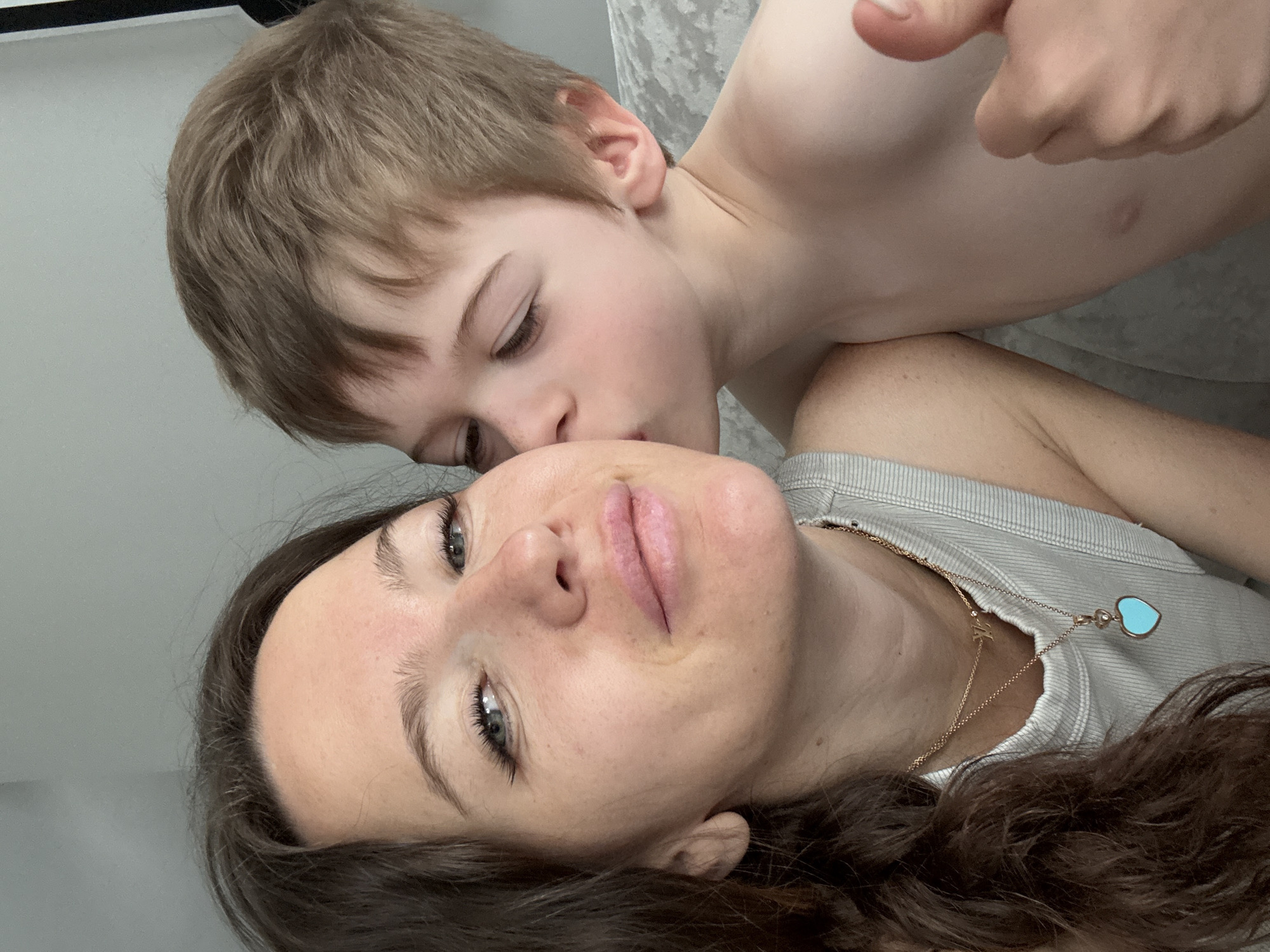 A close-up, angled shot shows a woman lying down while a young child leans over her, appearing to kiss her forehead. The woman has dark hair and fair skin, wearing a grey striped top with a gold necklace featuring a light blue heart-shaped pendant. The child, shirtless, has blonde hair and fair skin. His hand is partially visible near his head, and his eyes are closed, his face close to the woman's forehead. The background is a neutral grey, possibly a wall or bed linen. The location in Moscow, Russia is not determinable from the image.