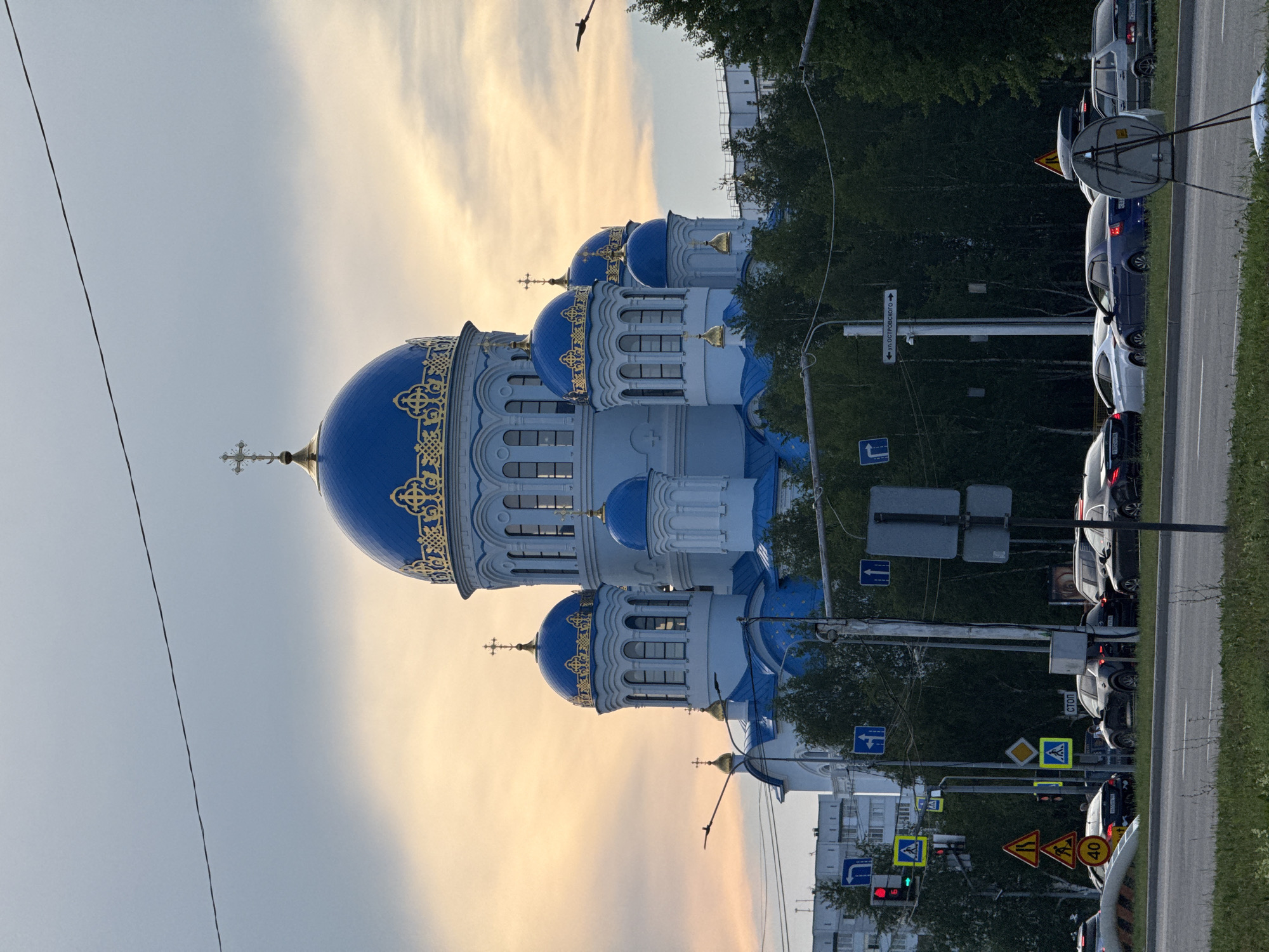This image captures a white and blue Orthodox church with golden ornamentation, featuring multiple domes topped with crosses. The church is situated near a road with several cars in traffic. The sky is a light gray and orange, indicating either sunrise or sunset. Lush green trees line the background, suggesting a natural setting within an urban or suburban area. Several road signs are visible, including traffic signals, speed limit signs (one indicating 40), and directional signs. One sign appears to read "ул. ОСТРОВСКОГО" which translates to "Ostrovskogo Street". The overall scene suggests a quiet moment during a transitional time of day, possibly dusk or dawn, with the traffic indicating normal daily activity. The lighting is soft, contributing to a serene atmosphere.
