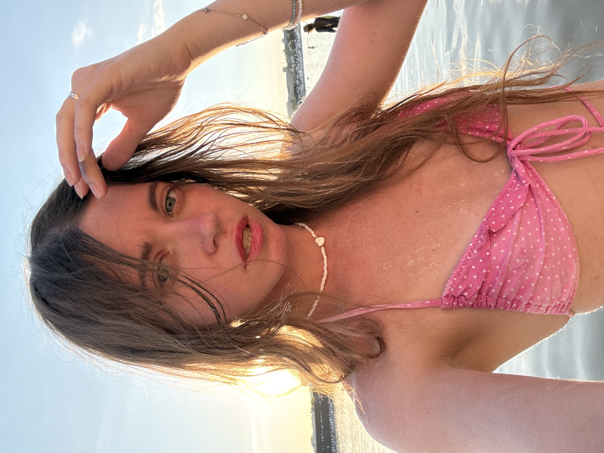 The media file is a close-up, rotated selfie of a young woman in a pink polka-dot bikini. She is in the water, with the sun setting behind her, casting a warm glow. The water is calm and appears to be shallow, with sunlight reflecting on its surface. The woman's hand is resting on her head, and her hair is wet and windswept. She is wearing a white beaded necklace with a small heart pendant. In the background, there is a faint outline of a city skyline and a person standing further in the water. The overall mood is relaxed and summery. The time of day is sunset, and the weather appears to be warm and clear. The location is likely a beach or a body of water in Sol-Iletsk, Russia, as suggested by the context. There is no visible text in the image.