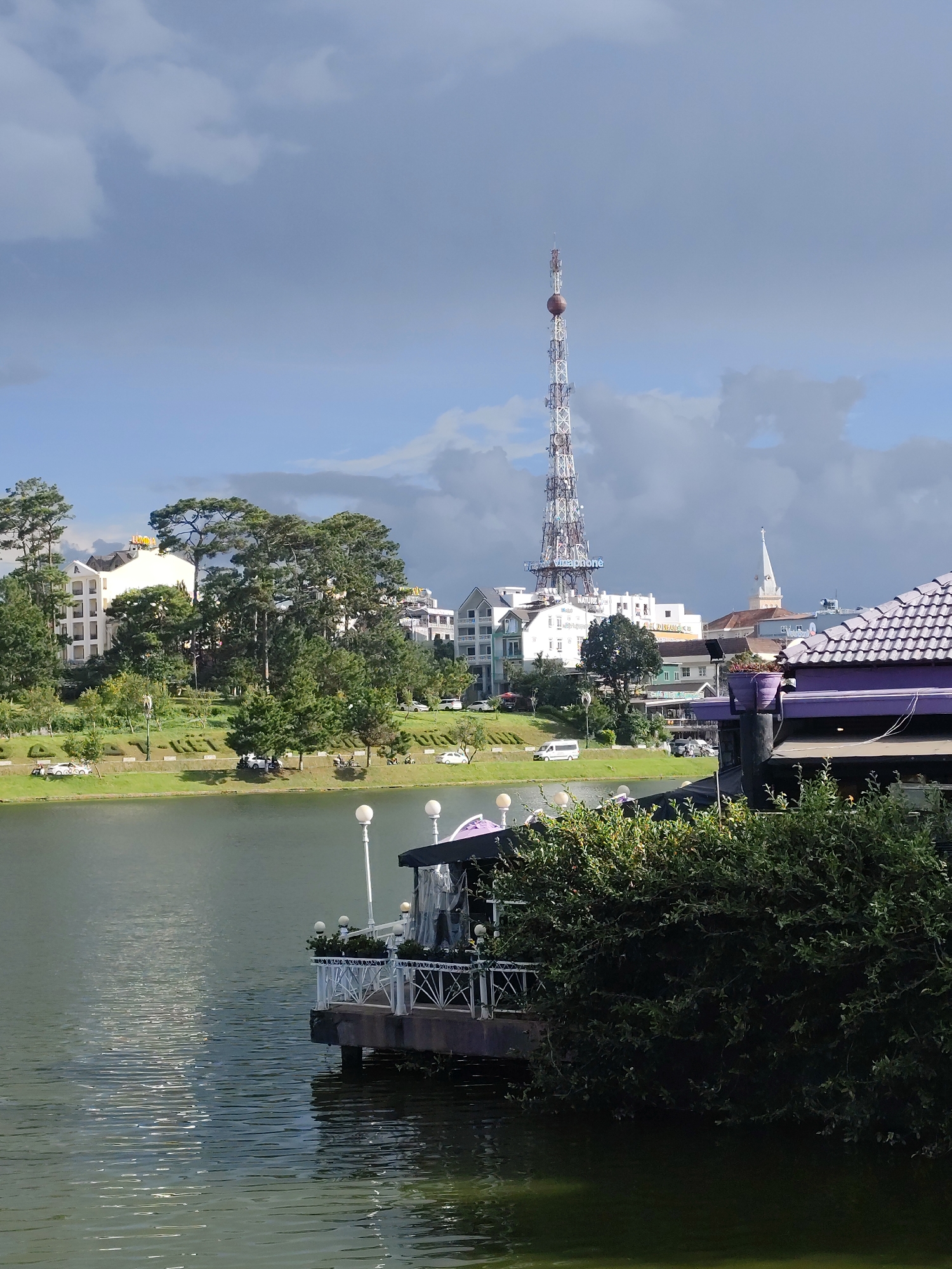 This is a scenic view of Xuan Huong Lake in Da Lat, Vietnam. The foreground features a portion of a building with a purple roof and a deck with white railings extending over the water. Lush green bushes are prominent on the right side of the foreground, partially obscuring the deck. The lake itself is calm, reflecting the sky and surrounding scenery. In the middle ground, a line of trees separates the lake from a collection of buildings. A prominent radio tower stands tall behind these buildings. The sky is partly cloudy, with dramatic dark clouds on the left and lighter, brighter clouds on the right. The buildings appear to be a mix of architectural styles, including a taller white building with a prominent sign that reads "LUMINA." Several cars are visible on a road running alongside the lake. The overall impression is that of a picturesque urban landscape adjacent to a natural body of water, possibly on a day with changing weather.