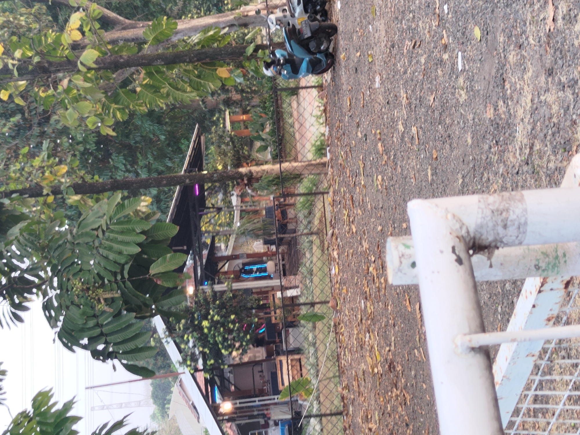 This image captures a daytime scene at what appears to be an outdoor cafe or restaurant in Depok, Indonesia. In the foreground, a weathered white metal fence and gate structure is visible, partially obstructing the view. The ground is covered with gravel and scattered fallen leaves.
Behind the fence, there's an open-air structure with a roof, likely a dining area, featuring wooden tables and chairs. Neon blue lights are visible inside this area, along with some illumination from warm-toned lights. A small tree with green fruits is situated near the structure.
In the mid-ground, on the right side of the image, a light blue scooter and a white scooter are parked. The blue scooter has a license plate. A chain-link fence separates the cafe area from the open space where the scooters are parked.
The background is dominated by lush green foliage from various trees, with some trees showing yellowing leaves. The sky is overcast, suggesting a cloudy day. Electrical pylons are faintly visible in the distance, indicating the proximity to urban infrastructure. There are no visible people in the image.