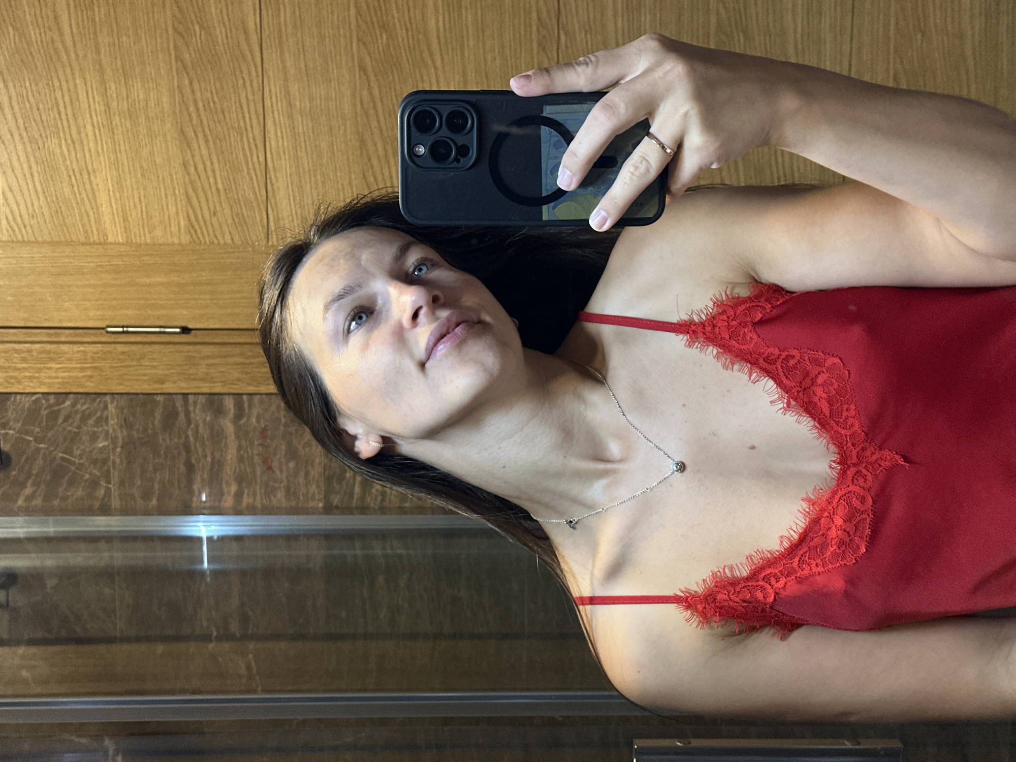 A woman is taking a selfie in a bathroom. She is wearing a red lace-trimmed camisole and a necklace. She is holding a black smartphone in her right hand, and a ring is visible on her ring finger. The background includes a glass shower door with marble tiling and a wooden cabinet. The location is a bathroom in Donji Pobori, Montenegro.