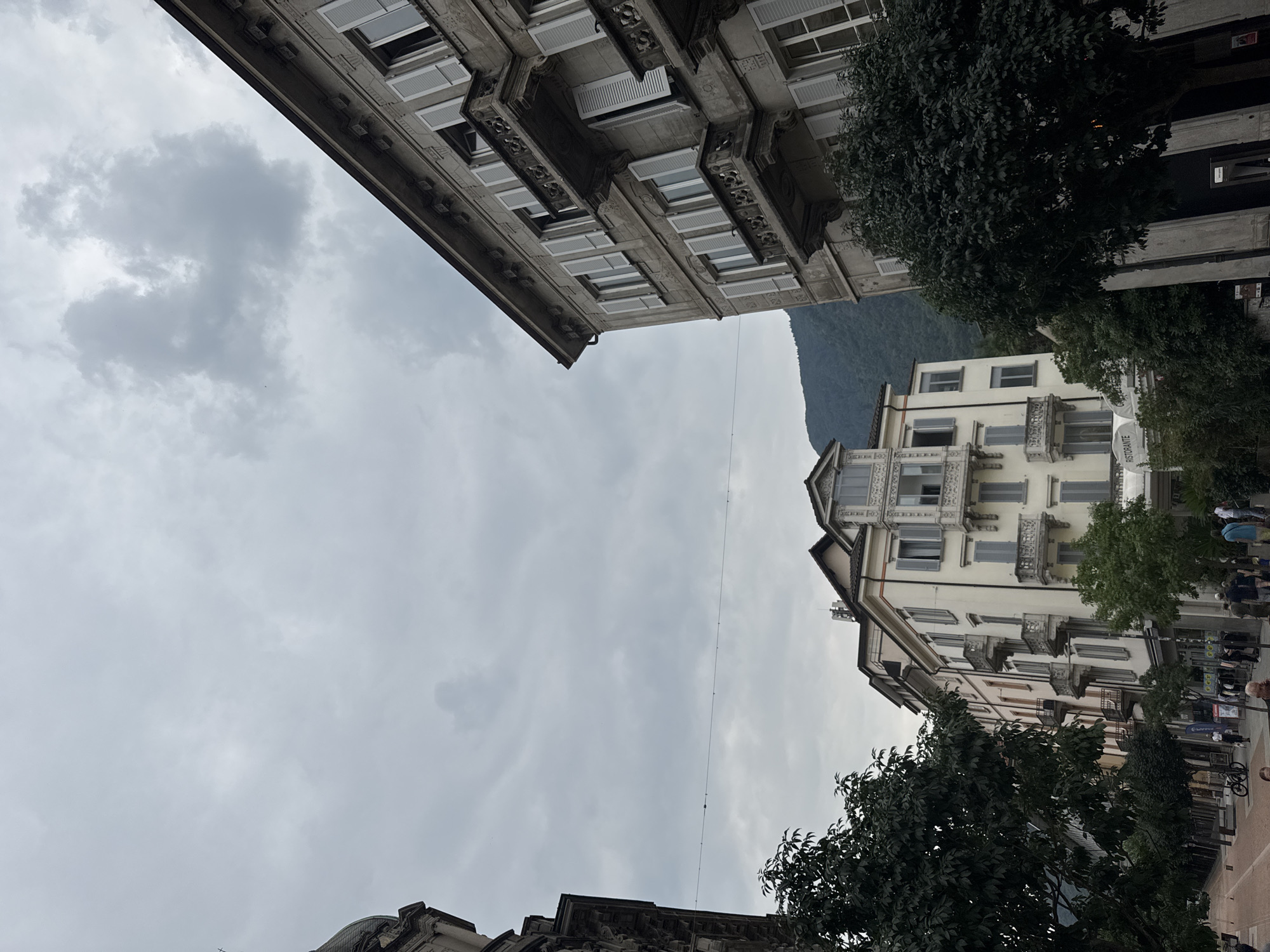 The image captures a view of buildings in Bellinzona, Switzerland, under a cloudy sky. In the foreground, a pedestrian walkway lined with trees leads to a street. Several multi-story buildings with traditional architecture are visible, featuring balconies and shutters. One building on the right has ornate detailing and balconies. To the left, a building with a dome-like structure is partially visible. People are seen walking on the street and sidewalk, some engaged in casual conversation. A bicycle is parked on the walkway. In the background, a lush green mountain rises, partially obscured by the buildings and trees. The overall scene depicts a typical European city street with a blend of historical architecture and natural landscape.