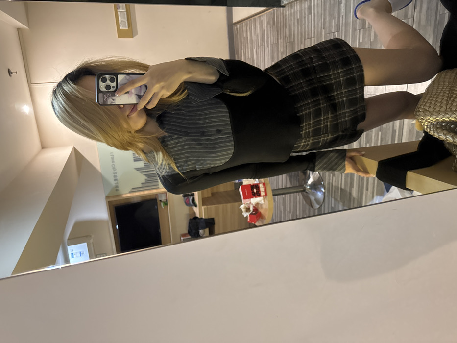 A person is depicted taking a mirror selfie in a room consistent with transient lodging, such as a hotel room, located in Yuzhong District, Chongqing, China. The individual, possessing blonde hair, is seen from the mid-thigh up, holding a black-cased smartphone featuring a light-colored graphic on its back, in their right hand to capture the image. Their face is partially obscured by the phone. They are dressed in a long-sleeved top with a grey and white striped collared upper section and a solid black lower section, paired with a dark brown plaid mini-skirt. Their left hand rests on a light-colored wooden furniture piece, while their right foot, wearing a light blue slipper, is slightly lifted.
The reflected background reveals a small, round table with a chrome pedestal base to the left, holding a red box and white tissues. Above the table, a black flat-screen television is mounted on a wooden shelf, with a red baseball cap and other dark items resting below it. An air conditioning unit is visible on the wall above the television. The wall features a mural with vertical black lines and mirrored Chinese text, partially decipherable as "I love Chongqing," confirming the location. The room has light-colored walls and a patterned floor resembling wood planks. A golden, woven bag is visible near the bottom of the reflection, positioned on the floor or furniture.