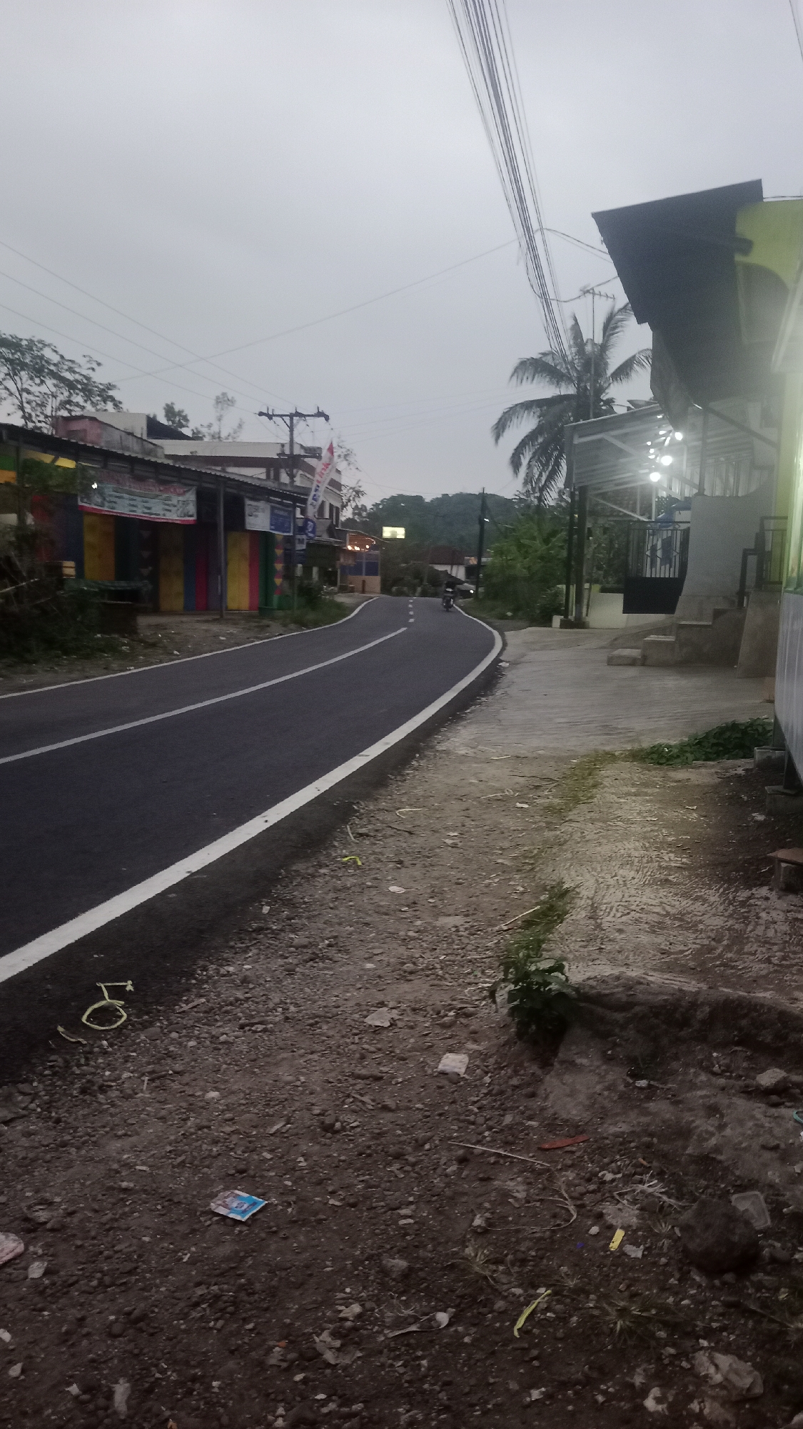 The image depicts a road in Cogreg, Indonesia. The road is paved and curves to the left. There are white lines marking the lanes of the road. On the left side of the road, there are several buildings with colorful facades. On the right side of the road, there is a sidewalk made of concrete. There are also some plants growing on the side of the road and sidewalk. In the distance, there is a person riding a motorcycle on the road. The sky is overcast, and the lighting suggests it is either early morning or late evening. The environment appears to be a semi-rural or suburban area with a mix of residential and commercial buildings.