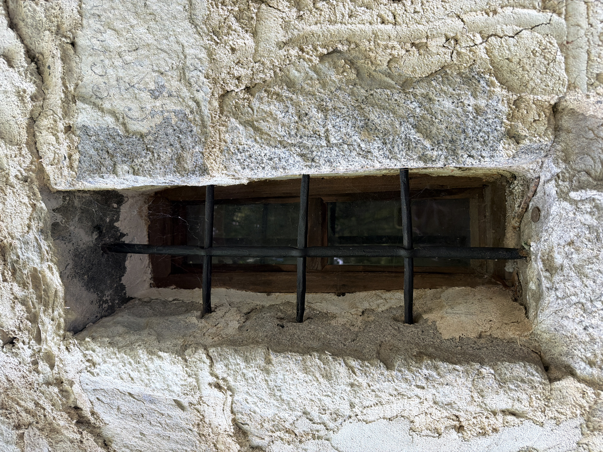 A barred window set into a rough stone wall is located in Govedartsi, Bulgaria. The window frame is made of wood and contains panes of glass. A grid of black metal bars covers the exterior opening. The stone wall appears old and weathered with textured surfaces. The light suggests it is daytime. No people are present.