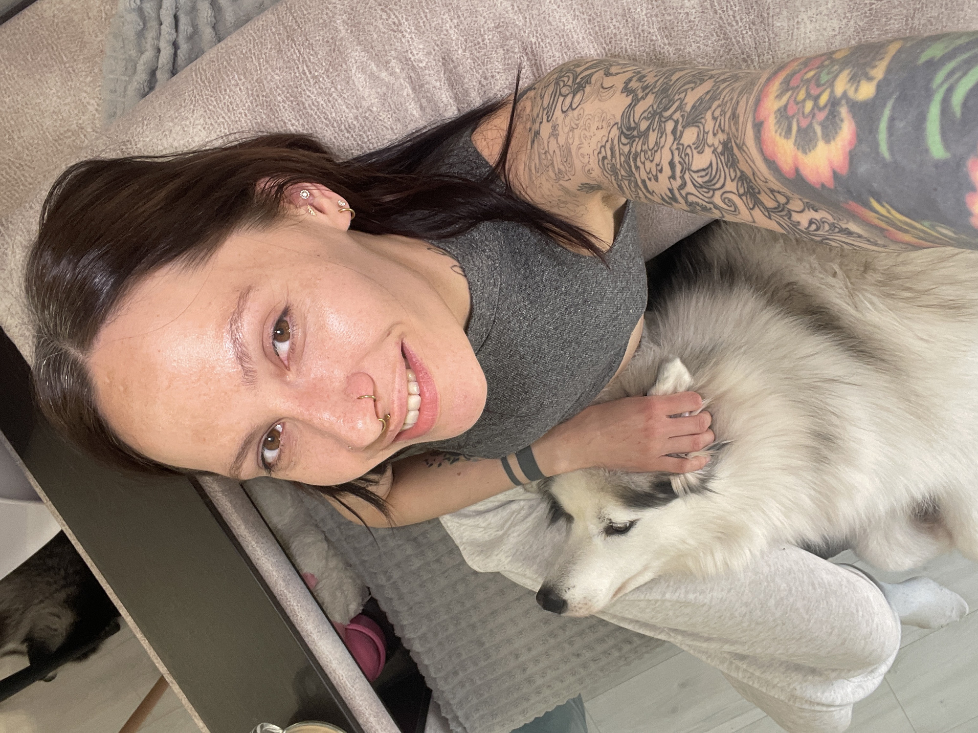 An overhead selfie captures a young woman and a husky dog in a relaxed indoor setting. The woman, with long, dark hair, is smiling warmly at the camera. She wears a grey tank top and is extensively tattooed; her right arm features a vibrant, colorful sleeve with red, orange, yellow, and green patterns, while her left arm displays black ink designs, including two solid dark bands on her forearm. She also has multiple facial and ear piercings, specifically a gold nostril ring, a septum ring, and various ear studs and hoops.
Her left hand gently rests on the back of a white and grey husky, which lies comfortably with its head on her leg or lap. They are positioned on a soft, light-colored, textured blanket or throw, likely on a sofa or bed. The setting appears to be a brightly lit interior, suggesting daytime. In the lower left corner of the image, part of a dark piece of furniture or shelf is visible, and a glimpse of a cat can be seen sitting nearby. The overall atmosphere conveys contentment and affection between the woman and her pets. There is no visible text in the image. The location context provided is Izhevsk, Russia, though this is not visually discernible from the image itself.