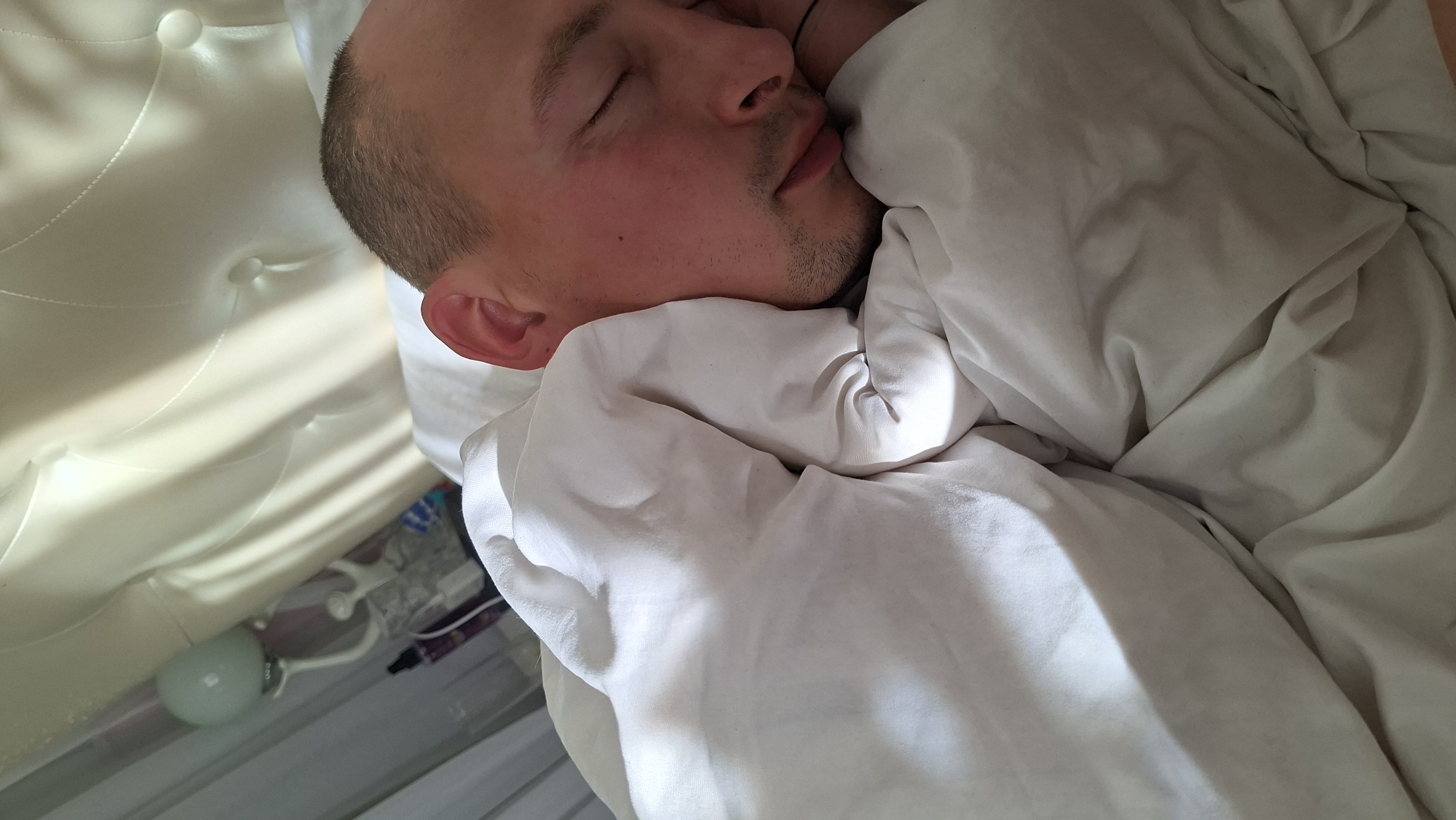 A man is sleeping in a bed, partially covered by a white comforter. He is lying on his side with his eyes closed, and his head is resting on a pillow. His skin is flushed red, and he has a light stubble on his chin. Behind him, a tufted headboard of a bed is visible, as well as a lamp with a light-colored shade. The scene appears to be indoors, likely in a bedroom.