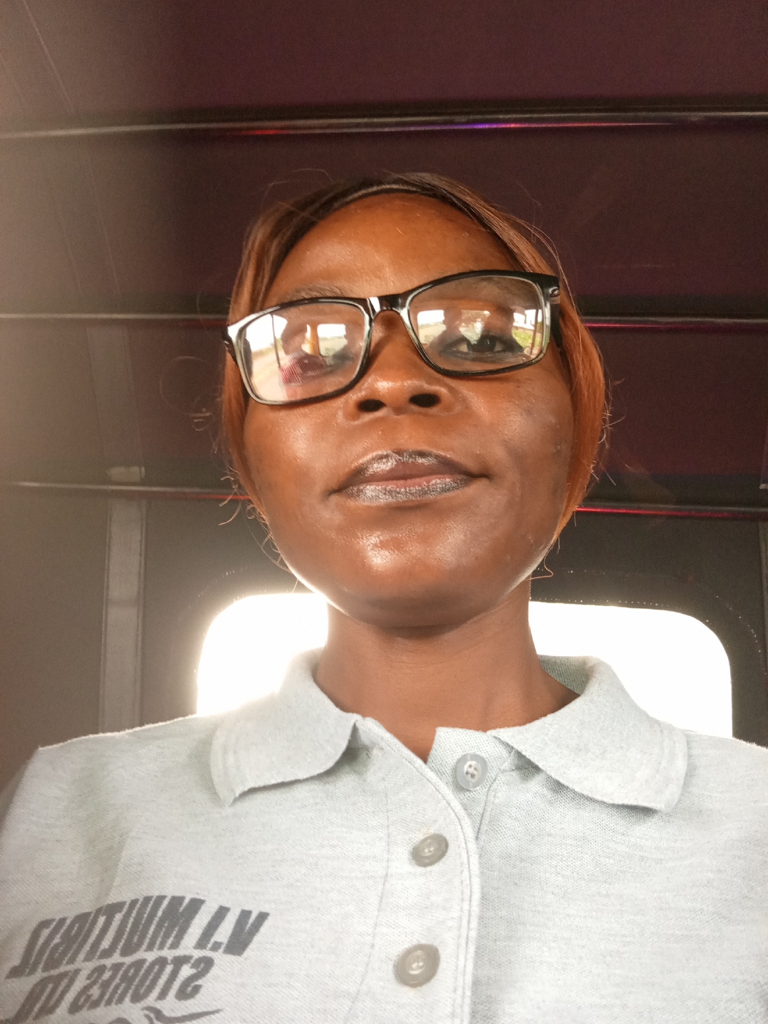 A woman is captured in a close-up selfie, seated inside a vehicle. She wears glasses and a gray polo shirt. The background suggests the interior of a tuk-tuk, with its characteristic overhead structure and dark, possibly vinyl, seating. The lighting indicates it is daytime, with a strong light source coming from the right, casting a bright glow. There is text on her shirt, partially obscured by the angle of the photo and the fabric's texture. The setting does not provide any specific contextual clues to place it in Mayo Dasa, Nigeria, or any other specific urban location.