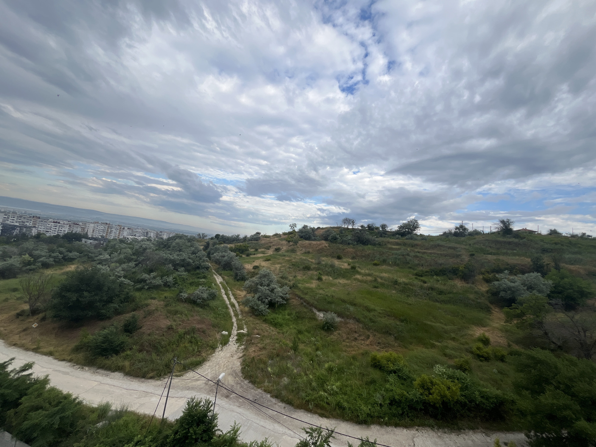 The image depicts a scenic view from a high vantage point overlooking the northern outskirts of Varna, Bulgaria, towards the Black Sea coast. In the foreground, a winding dirt road is visible, diverging from a paved road that leads into a dense green forest. The landscape transitions from lush vegetation to rolling hills dotted with scattered trees, some bare and others with sparse foliage, indicative of a steppe or scrubland environment. The horizon is lined with a cityscape, characterized by numerous apartment buildings, suggesting proximity to urban development. The sky is overcast with dramatic, textured clouds, allowing shafts of sunlight to intermittently illuminate the terrain. Several small birds can be seen in the sky. The overall impression is one of a natural landscape meeting urban expansion under a dynamic sky.