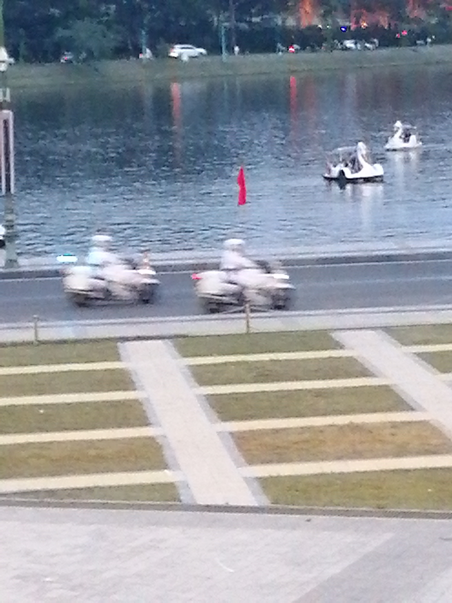 The image, taken in Da Lat, Vietnam, depicts a serene waterside scene. In the foreground, paved walkways and manicured grassy areas indicate a park or recreational space. Beyond this, a road runs parallel to a large body of water, likely Xuan Huong Lake. On the road, two individuals on motorized scooters are visible, each wearing white clothing and helmets. The scooters are white, with one appearing slightly ahead of the other. In the water, two white swan-shaped paddle boats are afloat. One paddle boat is closer to the viewer and appears to have a person inside. A small red flag is planted in the water to the left of this paddle boat. In the background, trees and distant lights suggest the presence of the city. The overall atmosphere is calm, with the water reflecting the subdued lighting of the late afternoon or early evening.