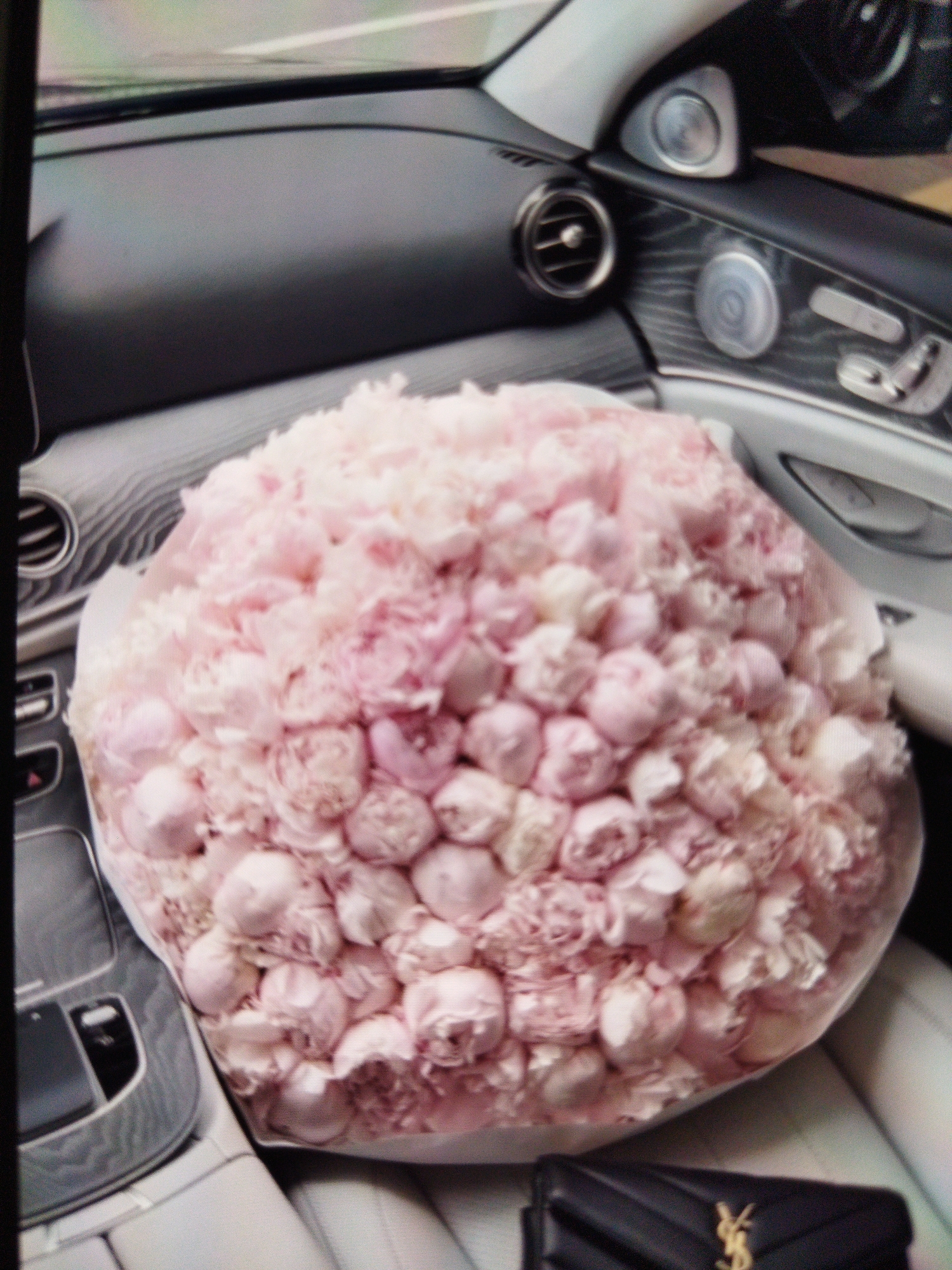 A large bouquet of light pink peonies is placed on the passenger seat of a car. The bouquet is wrapped in clear cellophane. The car's interior is visible, including the dashboard, air vents, and speaker grills. The seats appear to be made of light-colored leather. A black clutch with a gold logo is visible on the floor in front of the seat. The image is taken from a high angle, looking down at the bouquet. The lighting suggests it might be daytime, but there are no clear indicators of time of day or weather. The scene appears to be in a car, likely parked, as the focus is on the bouquet and interior. There is no visible text in the image.
