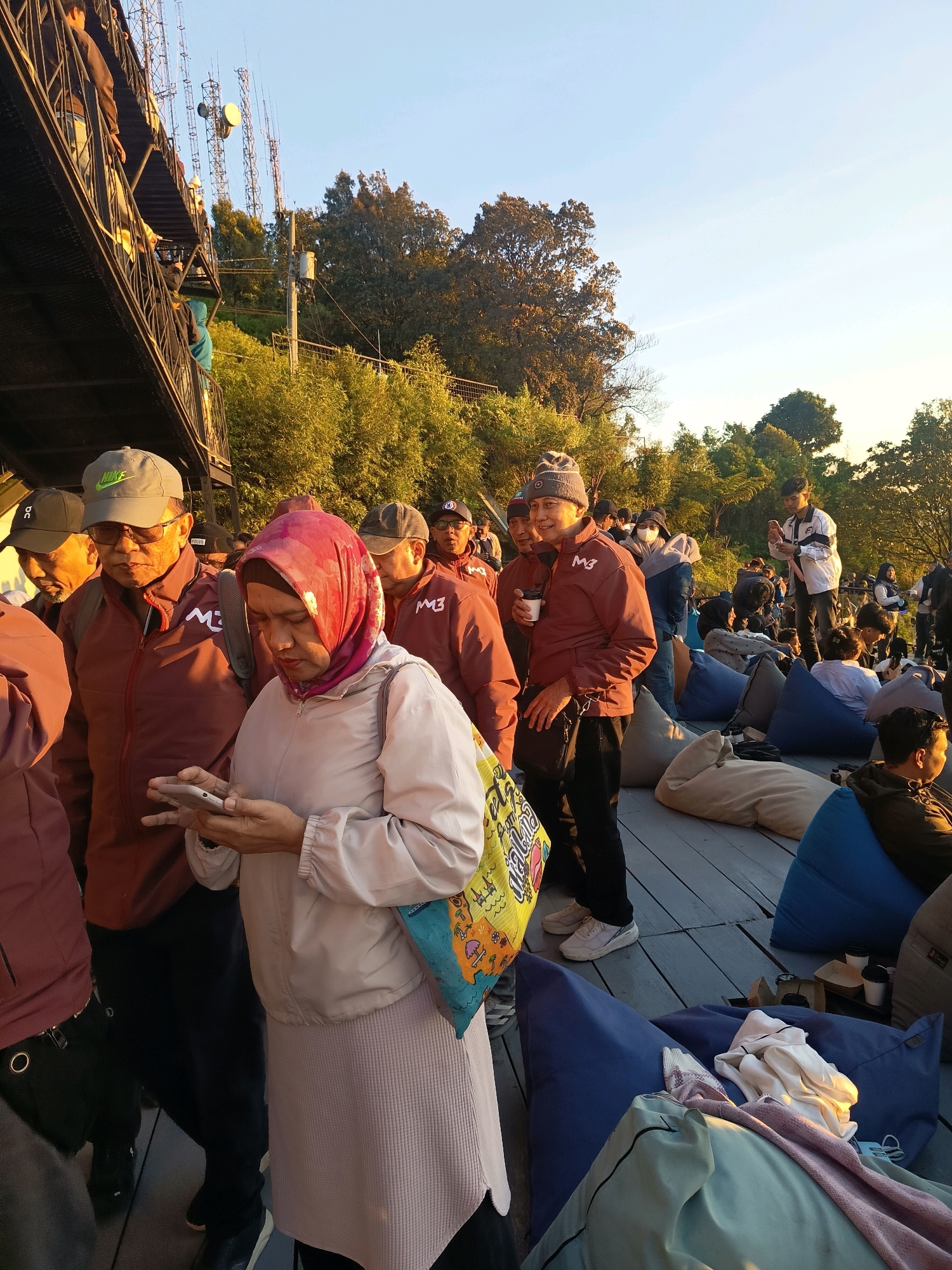 A group of people are gathered outdoors on a wooden platform, with some sitting on beanbag chairs. The scene appears to be in Keditan, Indonesia, judging by the style of the buildings and the vegetation. The lighting suggests it is late afternoon or early morning, with the sun casting long shadows.
In the foreground, a woman wearing a pink hijab and a light-colored jacket is looking at her phone. To her left, a man in a maroon jacket and a grey cap is also looking forward. Behind them, several other people are visible, some wearing similar maroon jackets with the "M3" logo. The background features trees and communication towers, indicating an elevated location.
Several beanbag chairs are scattered across the platform, some occupied by people. The overall atmosphere seems relaxed and casual, possibly a gathering or waiting area. The presence of communication towers suggests the location might be a viewpoint or a place with a good vantage point.