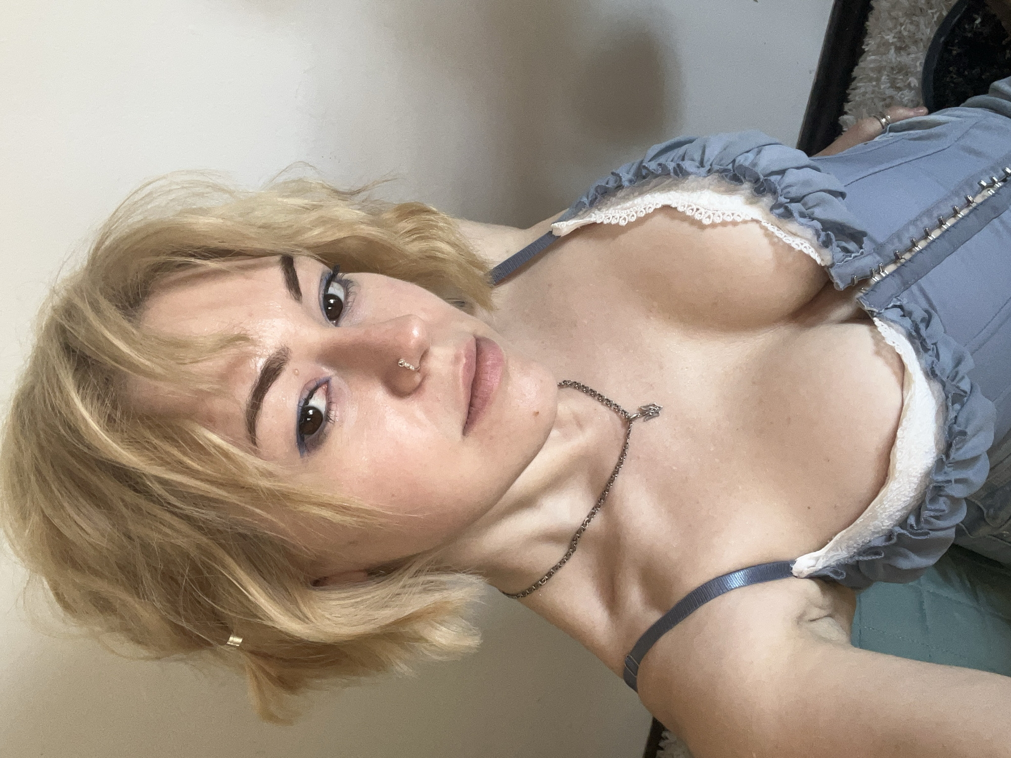 A female individual with short, blonde hair, wearing a light blue top trimmed with white lace and a silver necklace, is captured in a close-up, angled selfie. The scene is indoors, with a neutral-colored wall in the background and a hint of what appears to be a rug or dark object in the upper right corner. The image provides no visible information to indicate the location is Budva, Montenegro, or any city.