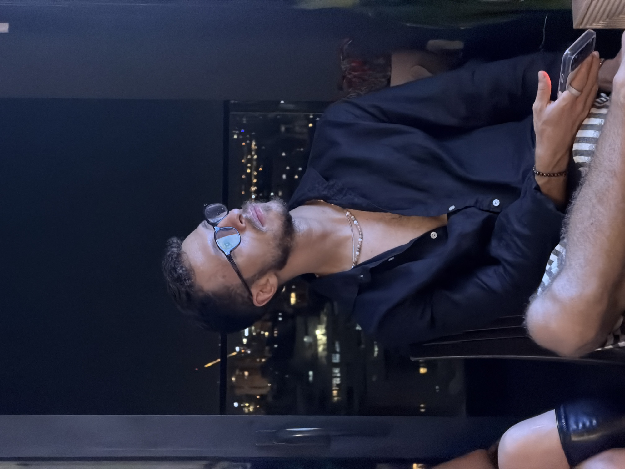 In Tel-Aviv, Israel, a young man with a beard and glasses is seated, looking to his right. He is wearing a black button-down shirt, open at the collar, revealing a beaded necklace. His lower half is clad in patterned shorts. In his right hand, he holds a transparent phone. The background suggests an urban setting at night, with city lights visible through a window, indicating the scene is likely indoors, possibly on a balcony or near a large window, overlooking the cityscape of Tel-Aviv.