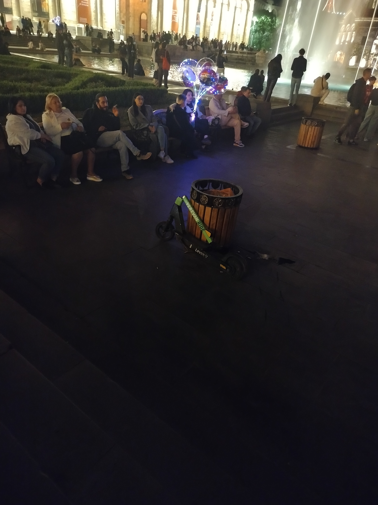 The image shows a night scene in Yerevan, Armenia, near the Yerevan Cascade complex. A group of people are seated on benches. The Cascade complex and its fountains are visible in the background. An electric scooter labeled "Yerevan Ride" is parked beside a trash can. There are pedestrians near the water fountain.