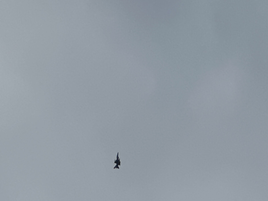 The image shows a single military aircraft flying in a clear, light gray sky. The aircraft is a dark silhouette against the sky, and its position and orientation indicate it is flying towards the right side of the image. The image was taken in the sky over Krushynka, Ukraine, at coordinates 50.235 latitude, 30.386 longitude. There are no people or other objects visible in the image.