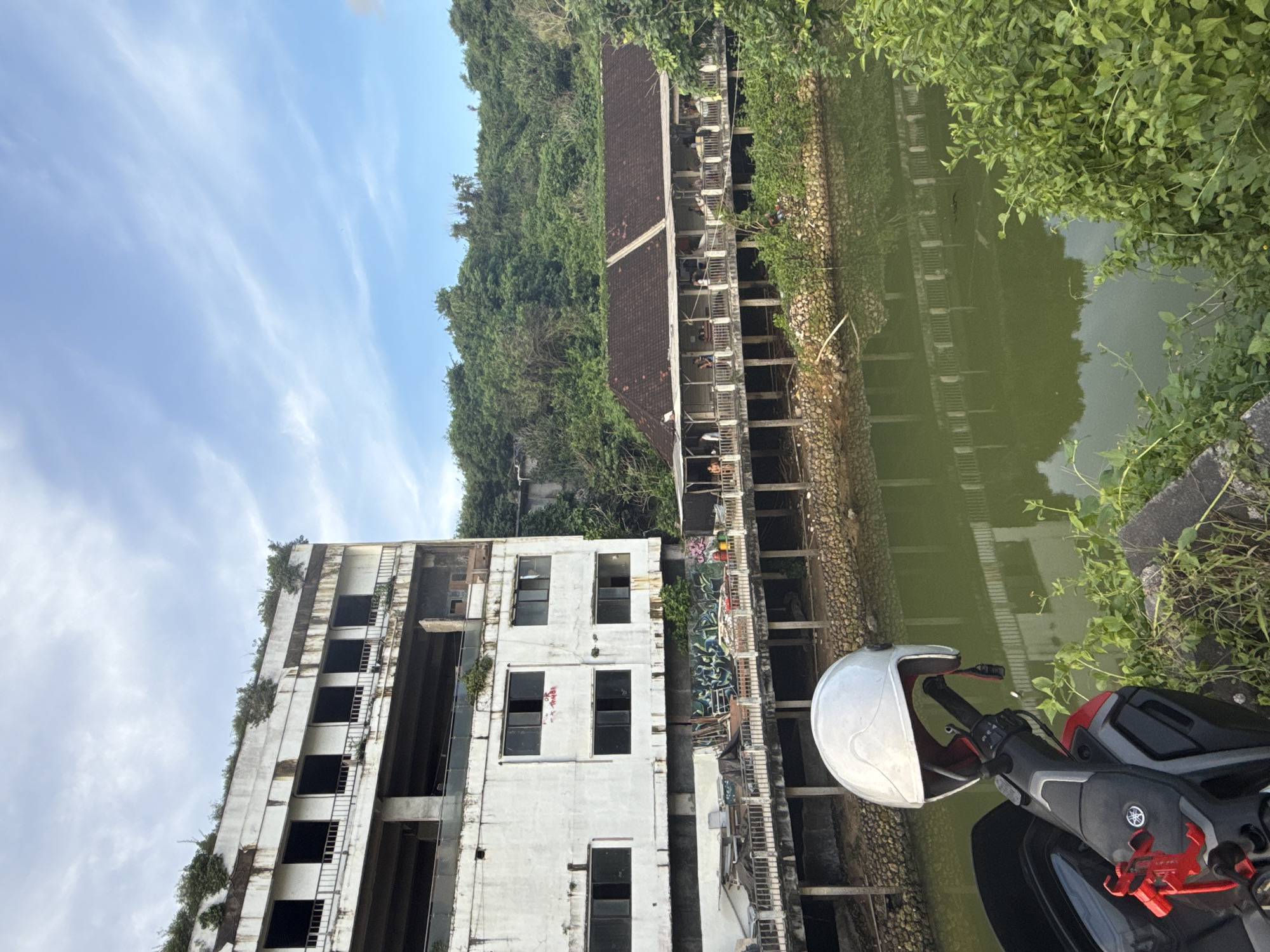 This image, taken in Kuta Selatan, Indonesia, presents an outdoor daytime scene featuring decaying urban architecture alongside lush natural elements. In the immediate foreground on the right, a white motorcycle helmet rests on the handlebars of a black and red scooter, which bears a visible Yamaha logo.
The central focus includes a large, multi-story white concrete building on the left, appearing significantly dilapidated and possibly abandoned. Many windows are dark and empty, and parts of the structure show signs of neglect, with vines and greenery beginning to grow on its ledges and roof. Graffiti, including an abstract blue and green design, is visible on its lower levels, along with some indistinct red text on an upper floor.
Separating this building from a green, forested hill on the right is a murky, green body of water, likely a pond or canal, featuring faint reflections. A textured stone wall borders the water near the white building. On the far side of the water, another multi-level structure with a dark tiled roof is visible, also appearing somewhat rundown and overgrown. Several people can be seen on its lower floors, suggesting partial use or access. The background behind this structure is a dense, vibrant green hill covered in trees.
The sky above is bright blue with scattered, wispy white clouds, indicating clear weather during the day. No specific activity or event is prominently featured, rather the image captures a static moment observing the environment.