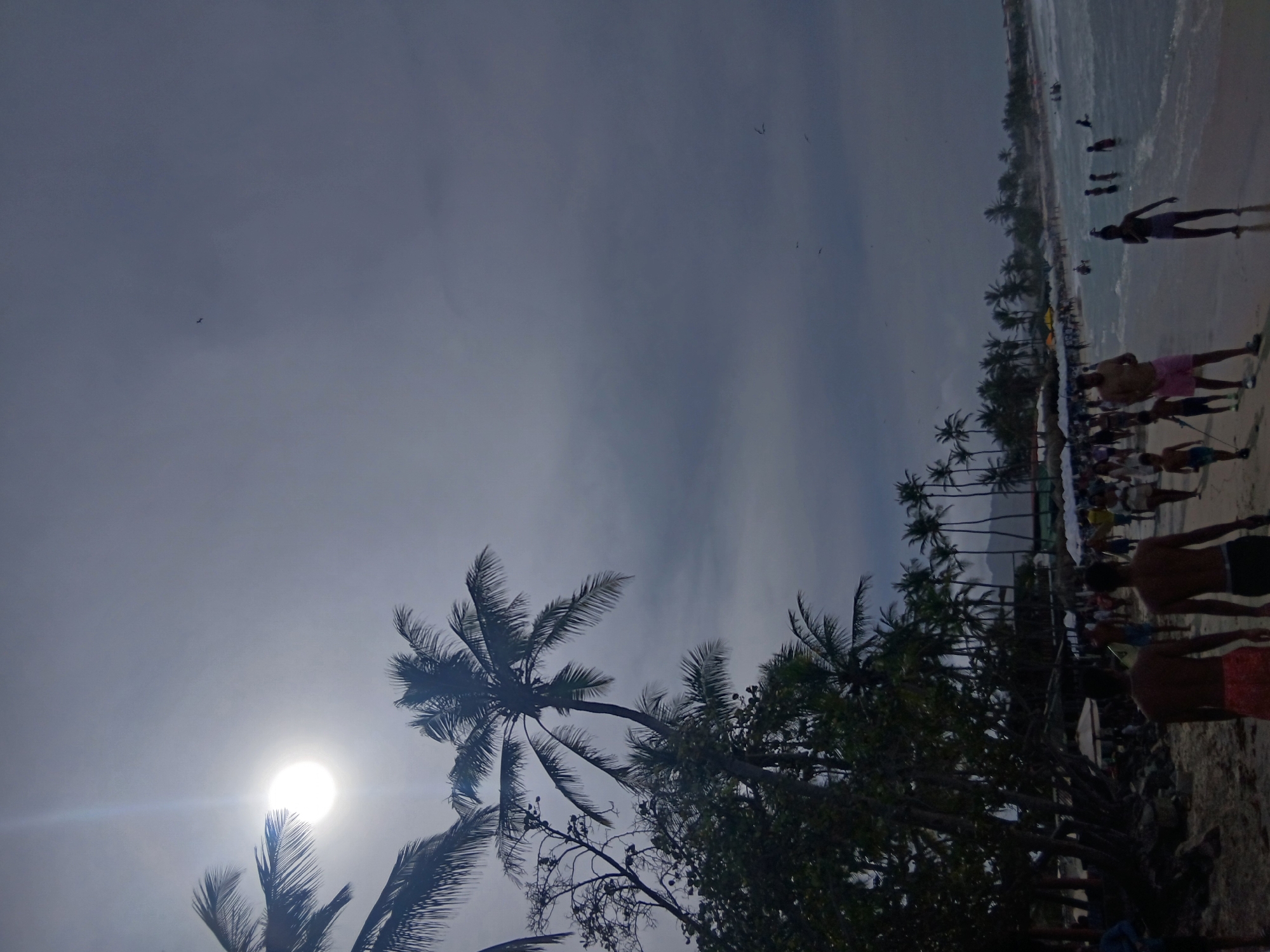 The image shows a beach scene at Playa Parguito, Venezuela. The sky is overcast, obscuring direct sunlight but the sun can be seen partially behind palm trees on the lower left. Palm trees line the beach separating it from the sand area where people are walking and standing. Several individuals can be seen wading in the water.
