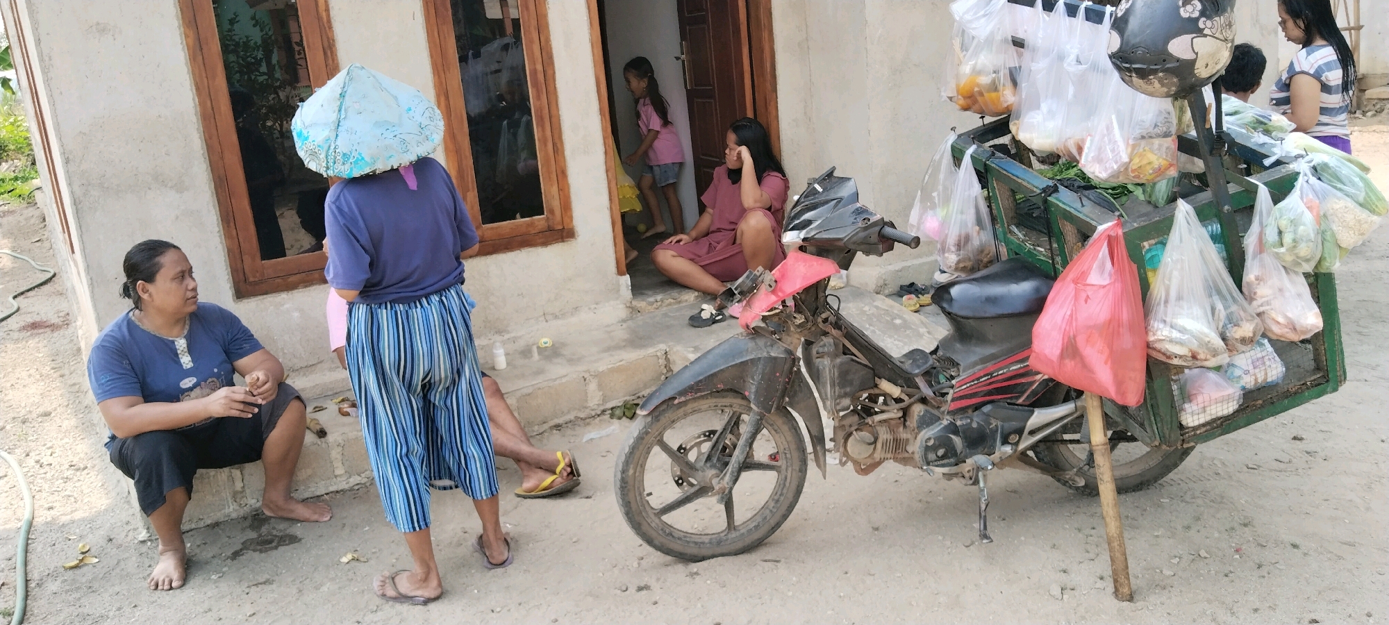 In Tanjung Ratu Ilir, Indonesia, a scene unfolds on a dusty, unpaved path beside a modest, light-colored dwelling with wooden-framed windows. A woman, with dark hair pulled back, is seated on the ground, wearing a navy blue t-shirt and dark shorts. She is holding a cigarette and looking to her left. To her right, another individual stands, wearing a navy blue t-shirt and striped pants, with a conical, light blue object on their head, possibly a hat or a basket.
A motorcycle, parked on the right side of the frame, serves as a mobile vending unit. It is fitted with a green metal rack containing numerous plastic bags filled with groceries. Visible items include what appear to be vegetables like cucumbers and leafy greens, as well as packaged goods and possibly fish. A red plastic bag hangs from a bamboo pole inserted into the ground next to the motorcycle.
Further into the doorway of the house, a young girl in a pink shirt and denim shorts is visible. Inside, another woman in a maroon dress is seated on the doorstep, looking away from the camera. The overall environment suggests a warm, possibly humid climate, with sparse vegetation and a general sense of everyday life in a semi-rural Indonesian setting. The ground is uneven and dry, with some debris scattered around.