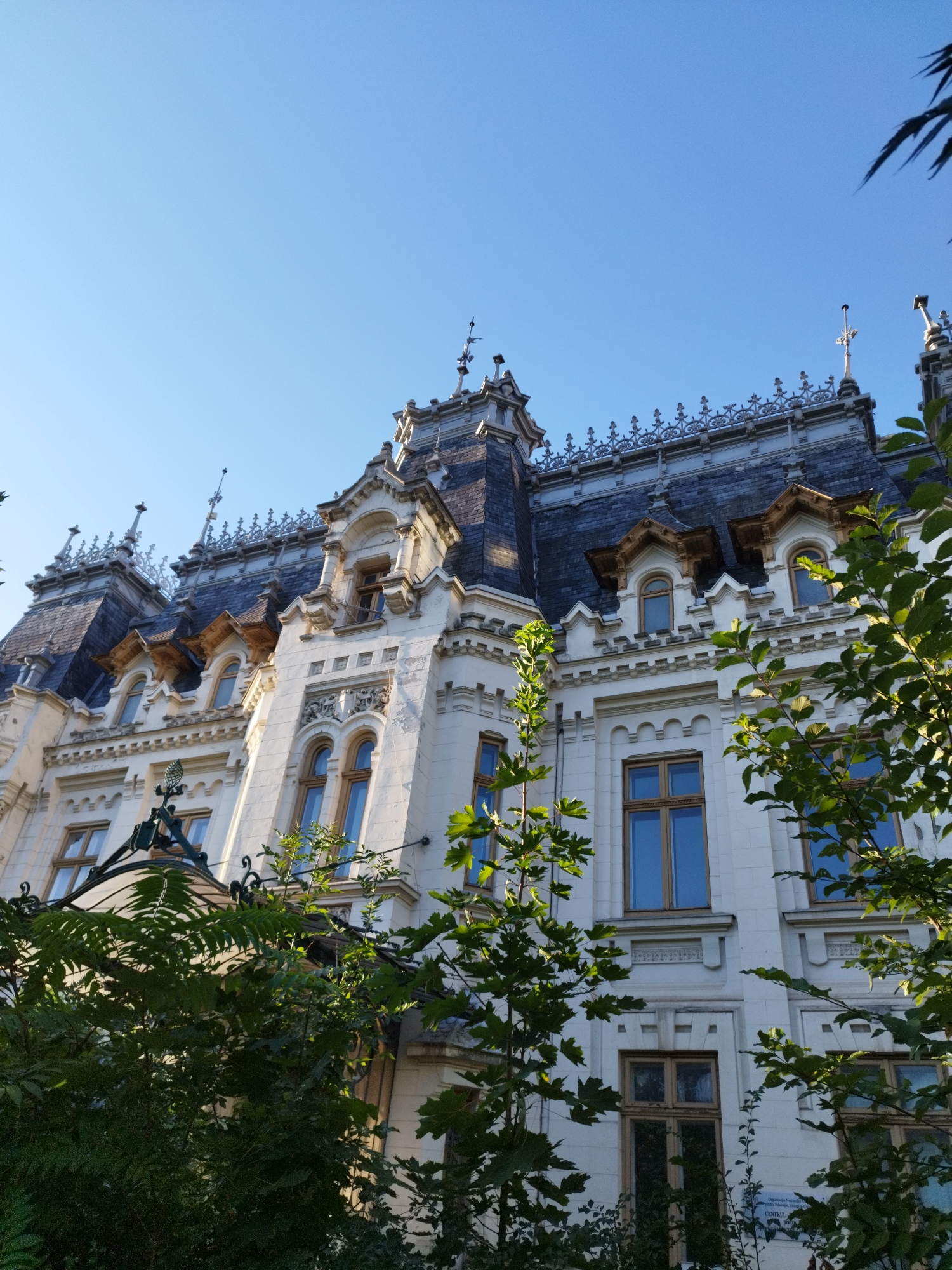 This is a low-angle shot of a grand white building with a dark, steeply pitched roof and ornate architectural details. The building appears to be a palace or a historic landmark, featuring multiple dormer windows with pointed roofs, intricate decorative moldings, and several spires topped with crosses. A wrought-iron fence or gate is partially visible in the foreground, and lush green foliage obscures parts of the lower facade. The sky is clear and blue. This building is the Cantacuzino Palace in Bucharest, Romania, located on Calea Victoriei. There are no people or animals visible in the image.
