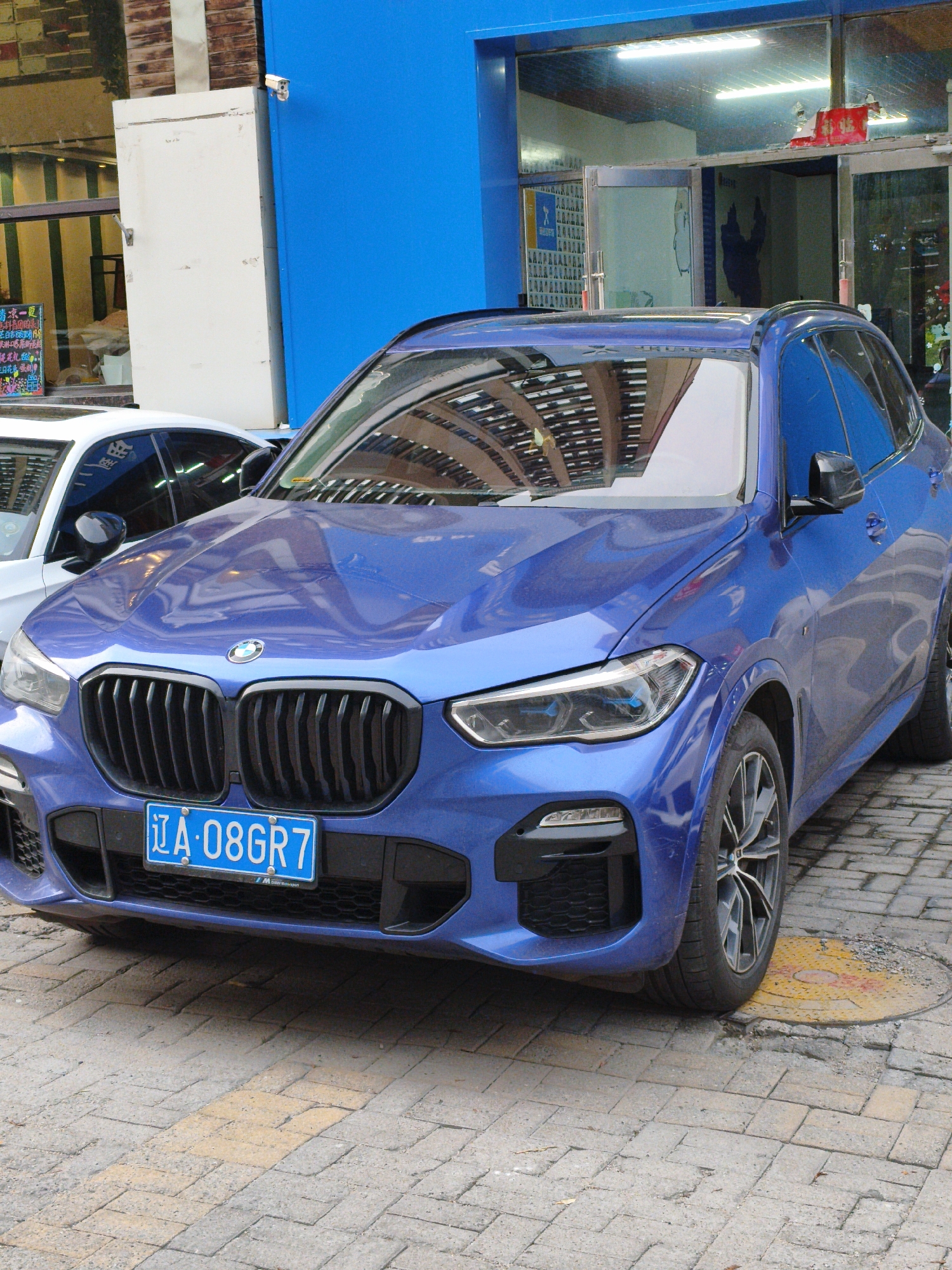 The image shows a blue BMW X5 parked on a brick-paved street in front of a building with a bright blue facade. The car is the primary subject, with its distinctive grille and headlights prominently featured. A white car is partially visible to the left of the BMW. The building behind the cars appears to be a commercial establishment, with a large window and a blue door. A security camera is mounted on the blue wall above and to the left of the white car. The scene is outdoors, with natural light suggesting it is daytime. The car's license plate is visible and reads "辽A·08GR7". There is also some Chinese text on a sign on the left side of the image, part of which is visible as "清凉一夏" and "三日花蛋糕". The street is paved with interlocking bricks, and a circular manhole cover with yellow markings is visible near the front right tire of the BMW. The overall impression is that of a car parked on a city street, possibly in a commercial area.