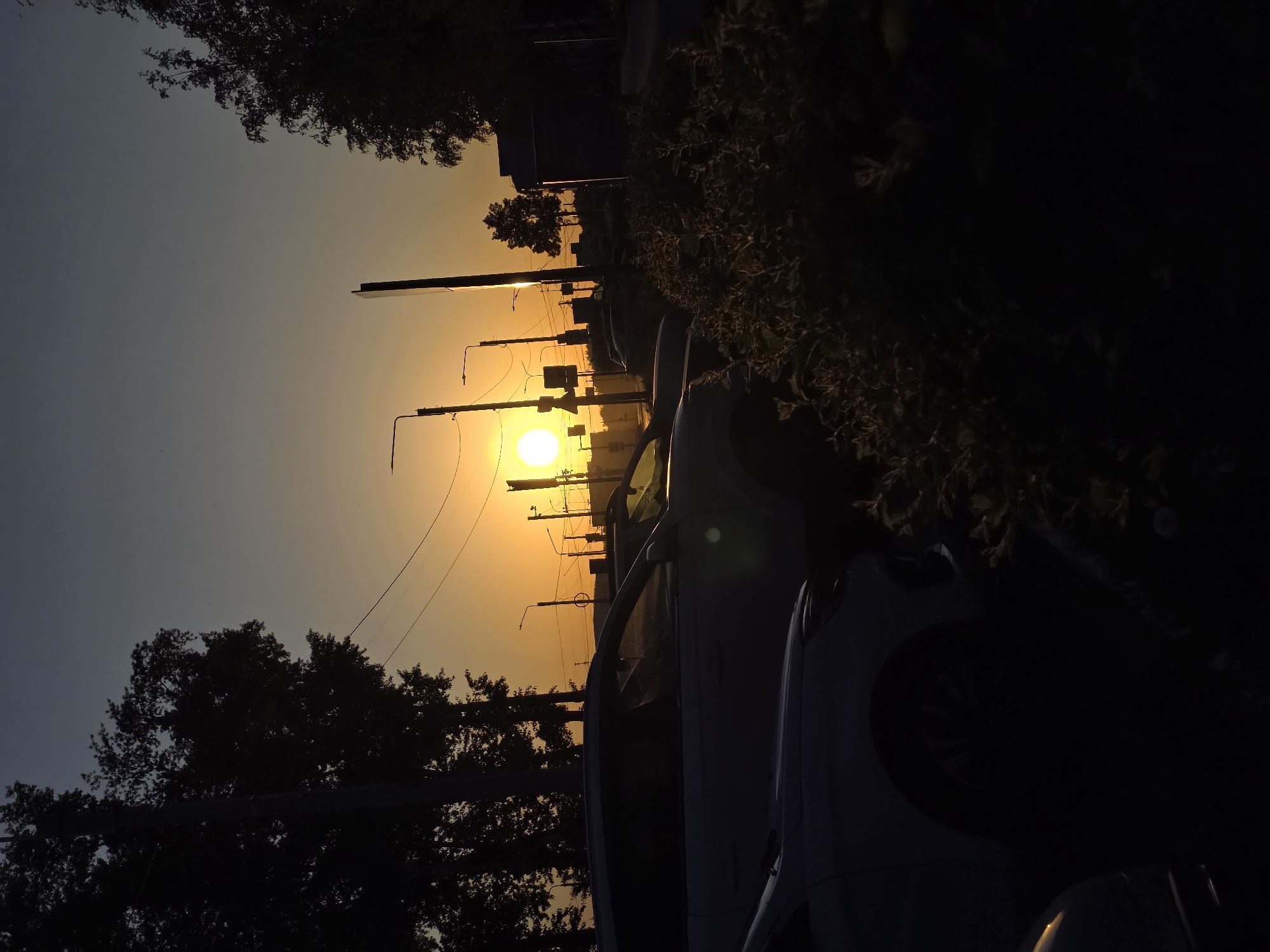The image shows a view facing east, with a setting sun. Cars are parked on a street on the right of the frame, silhouetted against the sun's glare. Overhead power lines stretch across the sky, connecting utility poles along the street. Trees are visible on either side, their branches silhouetted against the sky. The sky transitions from a dark blue-gray on the left to a bright yellow-orange gradient near the sun. This location could be in Chaikovsky, Russia, based on the presence of overhead power lines and the direction of the sunset. The photograph appears to be taken during the early evening hours.