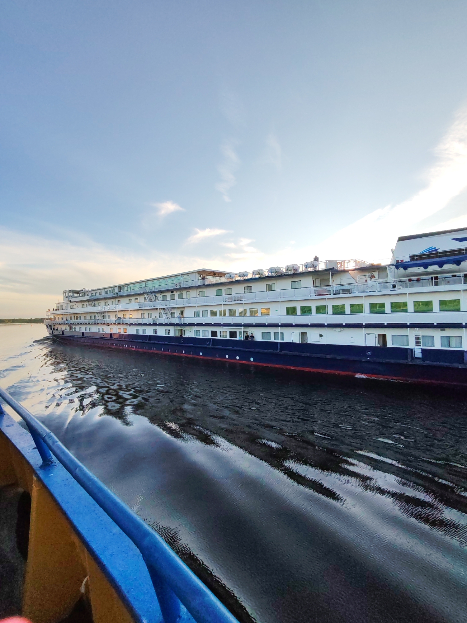 This media file captures a side view of a large, white cruise ship sailing on a body of water, likely a river, under a pale blue sky with scattered clouds. The ship, which appears to be a multi-decked vessel, has a dark blue hull with a red waterline. Several decks are visible, featuring numerous windows and balconies. Life rafts are stored on the top deck, along with some people who appear to be enjoying the scenery or engaged in conversation.
The foreground is dominated by the railing and part of the deck of a smaller vessel from which the photo is taken, indicated by the blue railing and a yellowish-brown surface. The water reflects the sky and the ship, creating a shimmering, textured surface. The overall lighting suggests it is either early morning or late afternoon, given the soft, diffused light and the clear sky with some wispy clouds.
The scene is serene and peaceful, conveying a sense of travel and leisure. The location is identified as Sheksna, Russia. While some text is visible on the side of the ship, it is not clearly legible in the provided image. The activity is the ship in transit on the water.