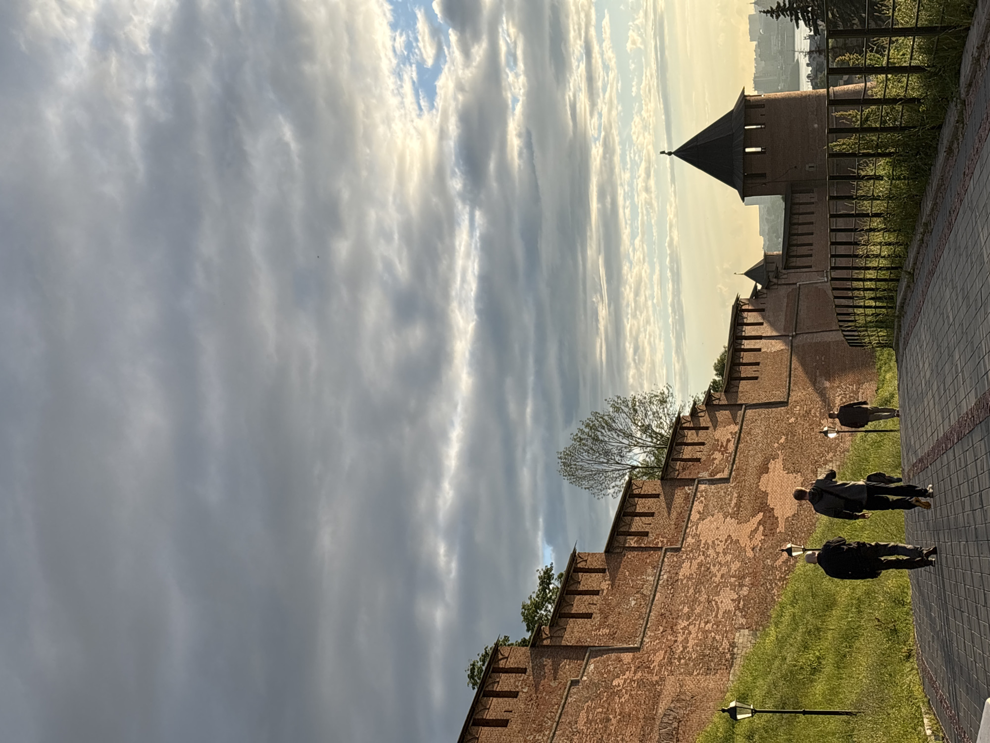 The image depicts a view of the Nizhny Novgorod Kremlin in Nizhny Novgorod, Russia, during what appears to be either sunrise or sunset. A red brick wall with crenellations runs diagonally across the frame, with a green grassy slope to its left. A paved walkway runs along the bottom right of the image, bordered by a black metal fence. Three men are walking away from the viewer on the walkway, two in the foreground and one further behind. A lamppost is visible on the left side of the walkway. In the background, a round tower with a dark conical roof, part of the Kremlin fortifications, stands prominently. Beyond the Kremlin, a cityscape with buildings and a body of water can be seen under a sky filled with dramatic, layered clouds. The lighting suggests a time of day with golden light breaking through the clouds, casting shadows and highlighting the textures of the brickwork and grass.