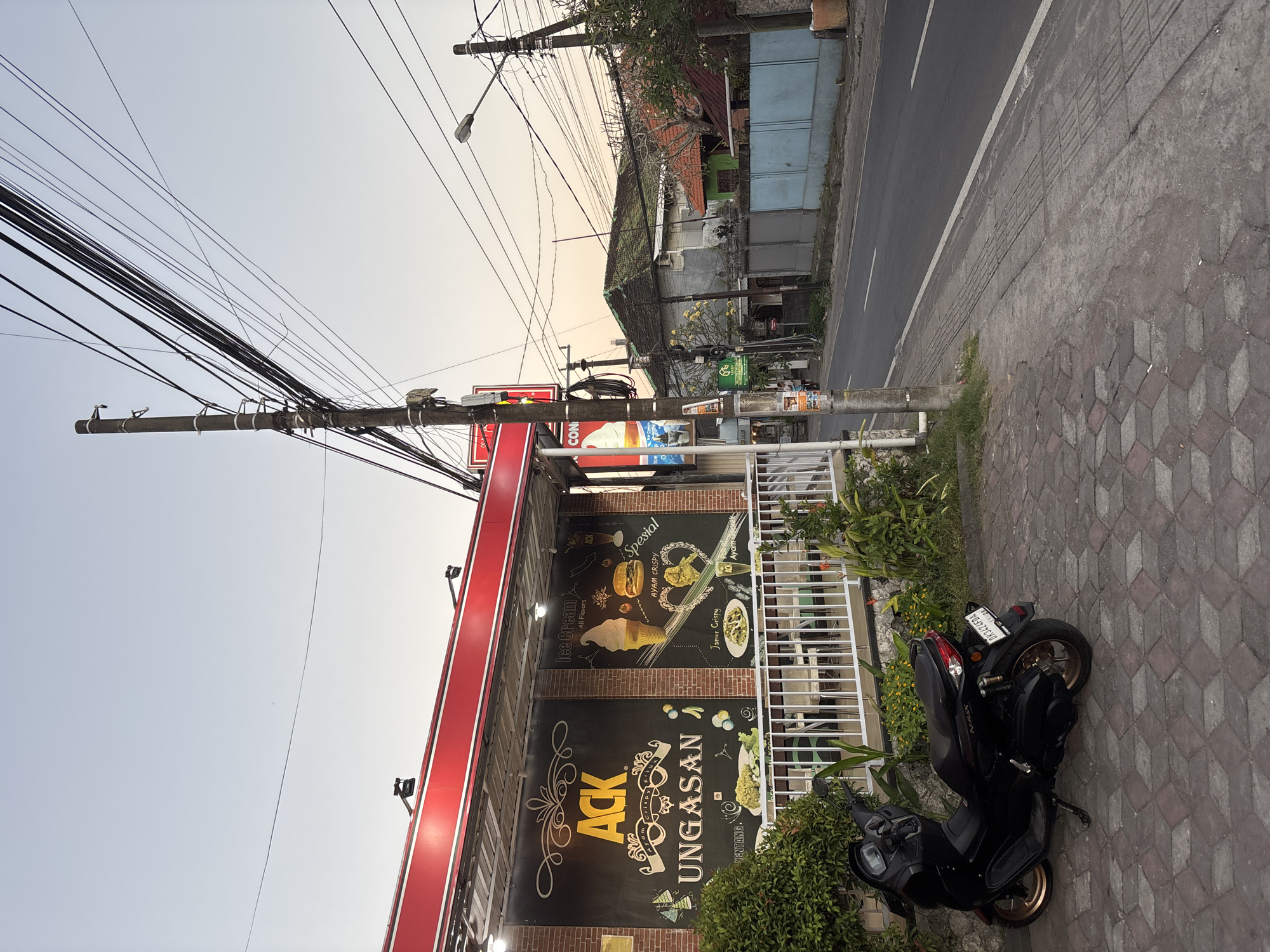 The image depicts a street scene in Kuta Selatan, Indonesia. A black motorcycle is parked on a patterned stone sidewalk in front of a building. The building has a red awning and large signs advertising "ACK UNGASAN," featuring images of food and ice cream. Utility poles with numerous wires are visible overhead. Across the street is another building with a light blue exterior. The sky is bright and clear.