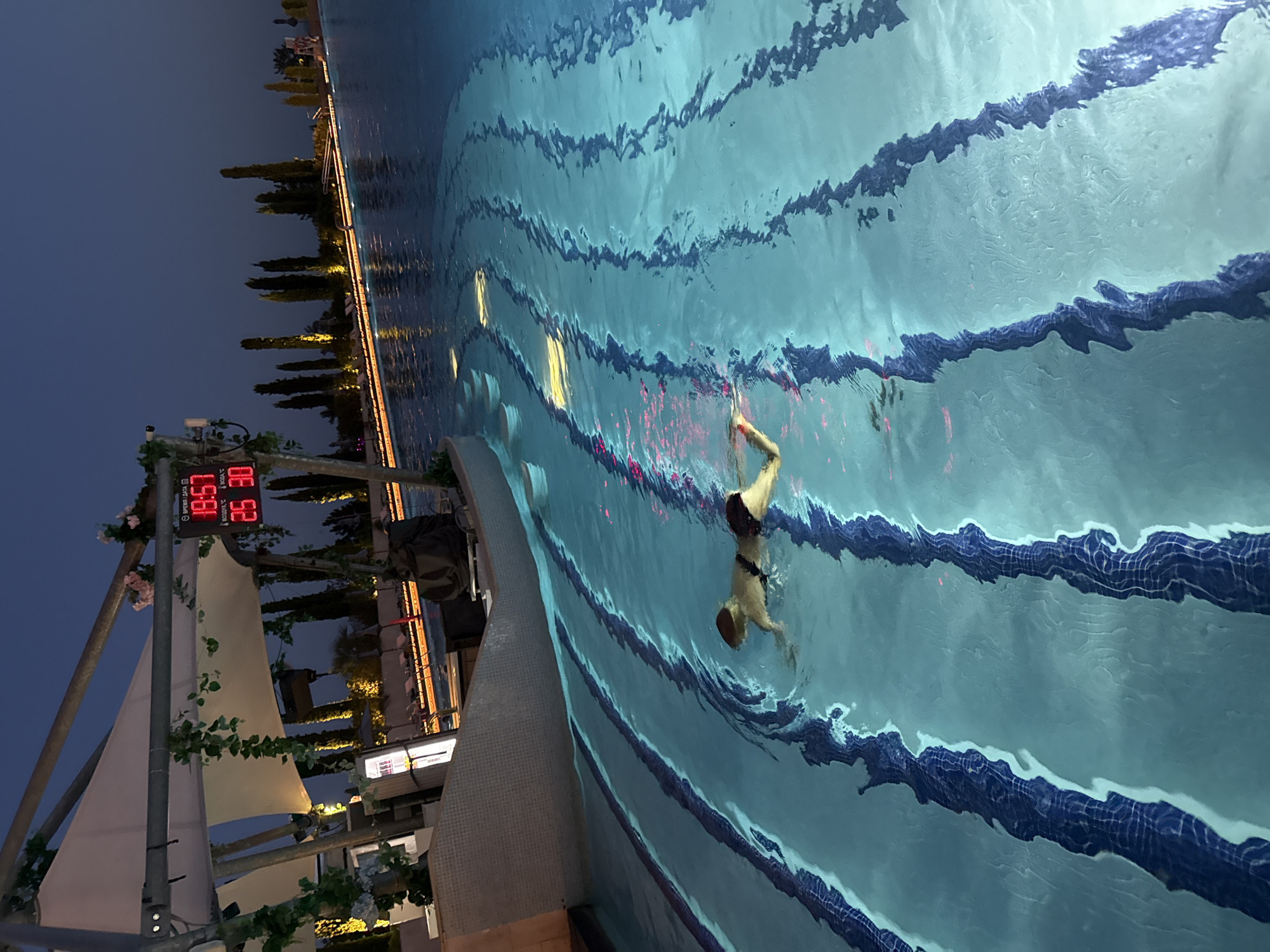 An outdoor swimming pool at night, with blue lane markers. Two people are swimming in the pool. A digital clock on the left side of the image displays the time as 19:57, the date as 25.30, and the water temperature as 30°C. The pool is surrounded by trees and lounge chairs. The location is Симеизкий поселковый совет, Ukraine.