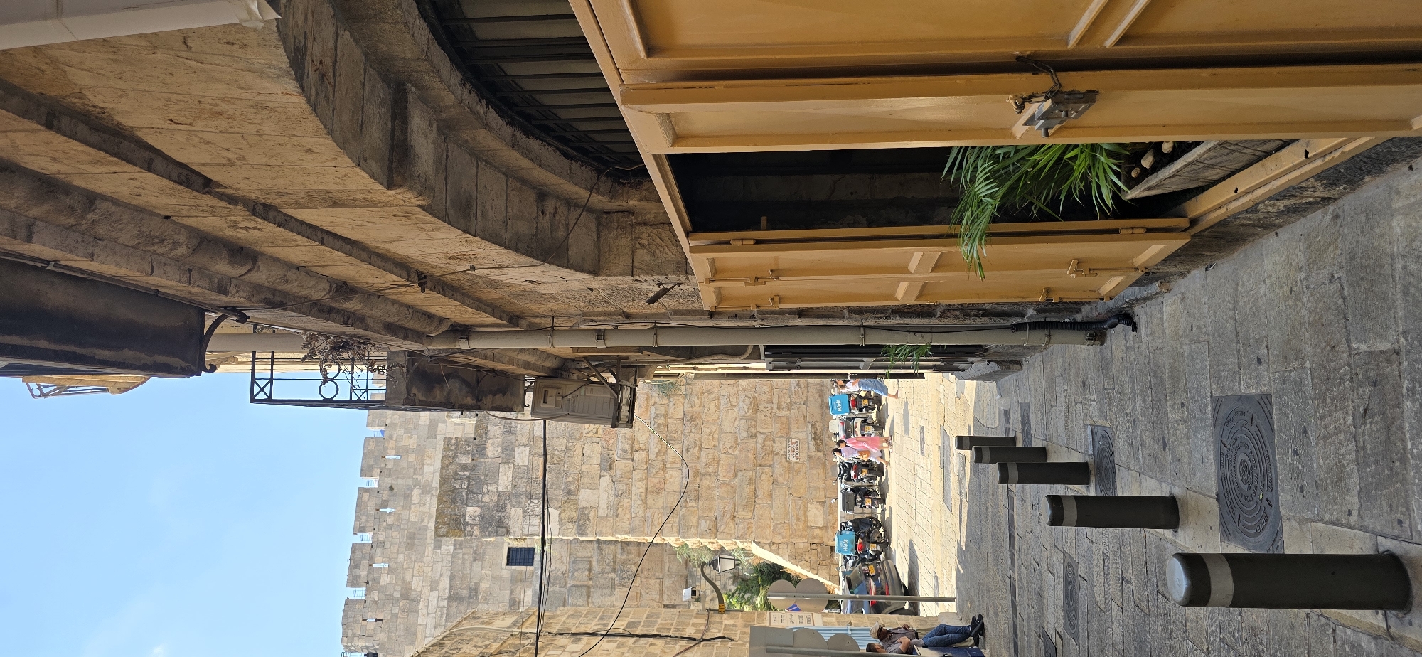 This is a vertical, medium-distance, eye-level, outdoor shot of a narrow street in the Old City of Jerusalem, Israel. The street is paved with stone slabs, with several dark gray bollards lining the right side of the walkway. On the left, a large, ancient stone wall with battlements and a small dark window is visible, believed to be part of the Old City walls. Further down the street, a row of scooters and two cars are parked. Several people are visible, including a man sitting on a bench to the far left, and three women walking past the parked scooters. The building on the right has a rough, stone facade and a large, open yellow door revealing a dark interior with a potted plant. The sky above is clear and blue.