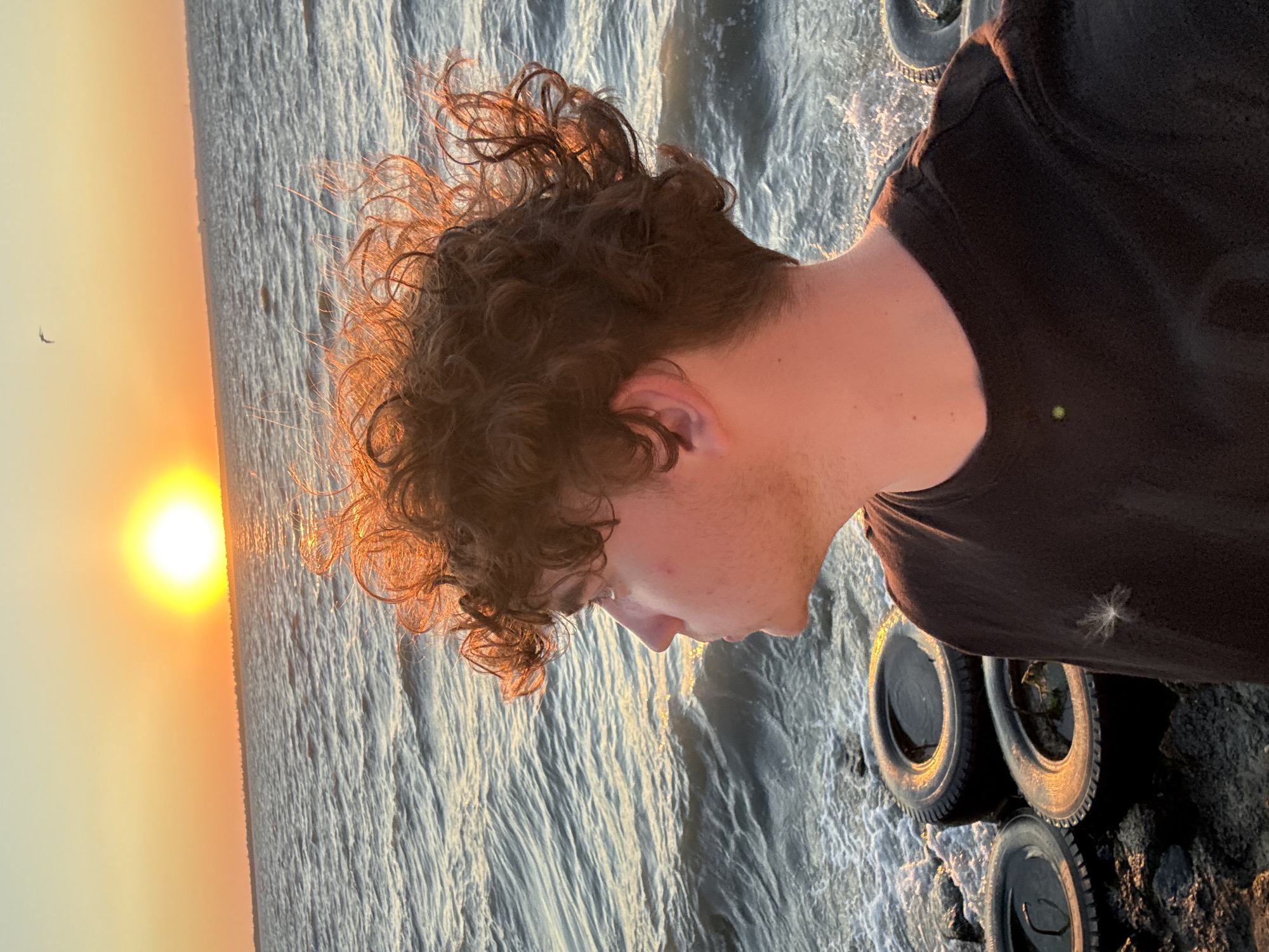 The image depicts a young person with curly brown hair standing on a rocky shore by the sea. The sun is setting over the water, casting a warm orange glow. In the foreground, several old tires are visible, some filled with water. The person is wearing a dark t-shirt and is looking out at the sea. The location is the city of Zatoka, Ukraine.