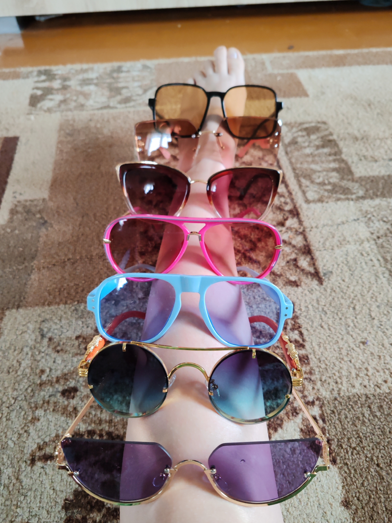 The image shows seven pairs of sunglasses arranged in a row on a person's leg. The sunglasses are of various styles and colors, including black, brown, pink, blue, and purple. The person's foot is visible at the top of the image. The background is a carpeted floor. There is no information available in the image to confirm or deny the location of the image as being in the city of Чухлэм, Russia.
