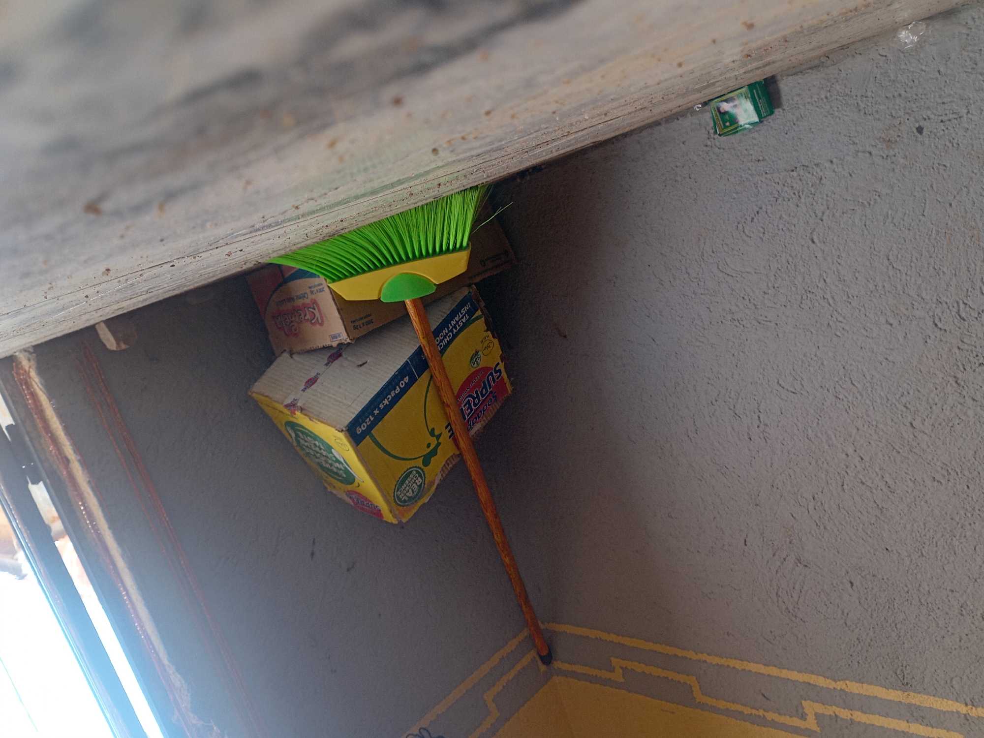 The image depicts an outdoor scene in Barakin Angundi, Nigeria, likely in a back alley or storage area. A green broom with a yellow handle rests against a textured gray wall, leaning against stacked cardboard boxes. One prominent yellow box features an image of a chicken and text indicating "REAL CHICKEN SOURCE" and "INSTANT NOODLE." Adjacent to it is a smaller box labeled "Kremela." In the lower left corner of the image, a small green and white package is visible on the ground, along with some scattered debris. The ground appears to be made of concrete or a similar material. The lighting suggests it is daytime, with sunlight entering from the right side of the frame, creating strong shadows and highlights. The overall environment appears somewhat weathered and utilitarian.